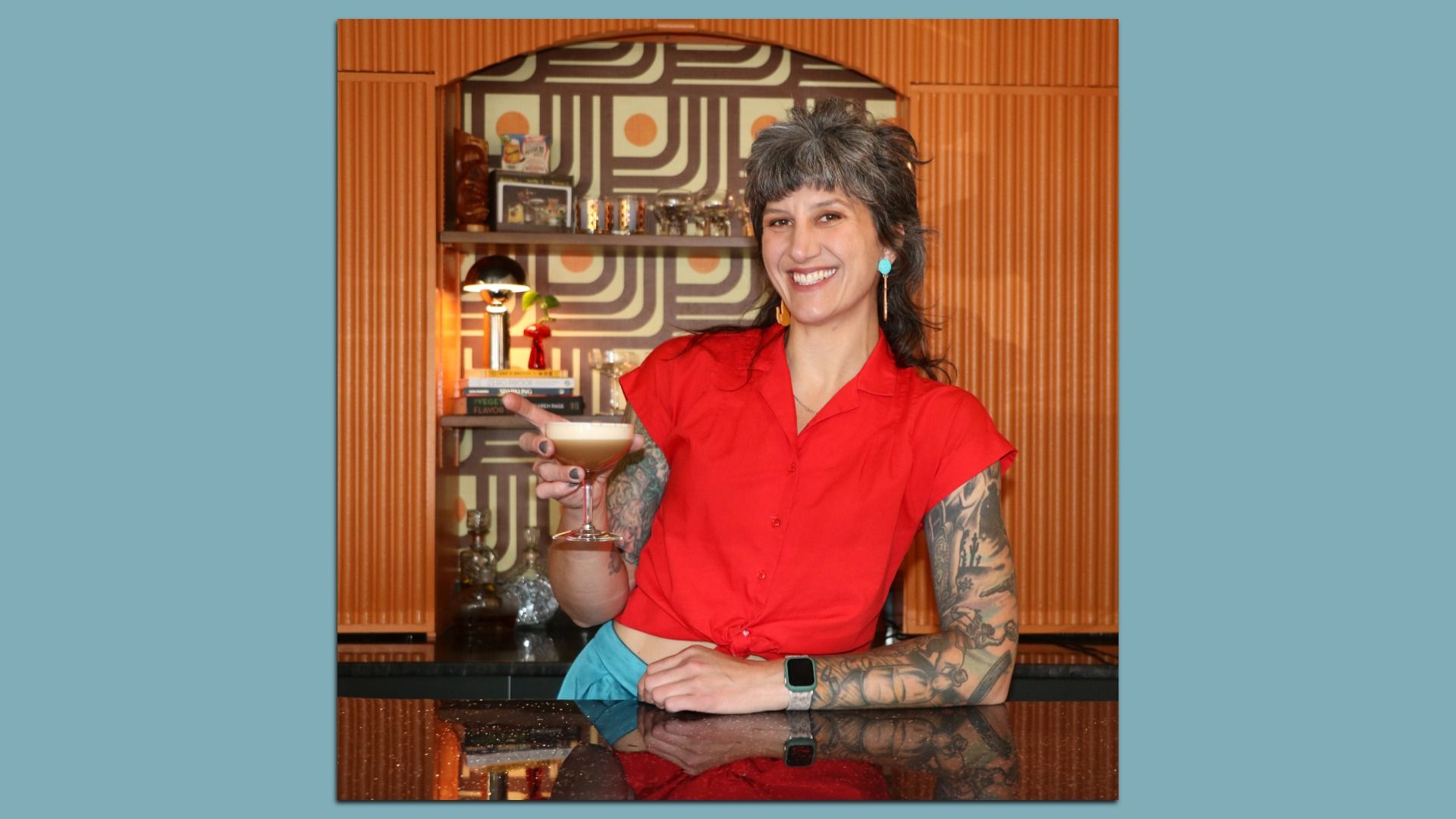 Smiling woman with short hair and tattoos on her arms, wearing a red blouse and turquoise earrings, holds a cocktail at a bar. Behind her is retro geometric wallpaper and shelves.