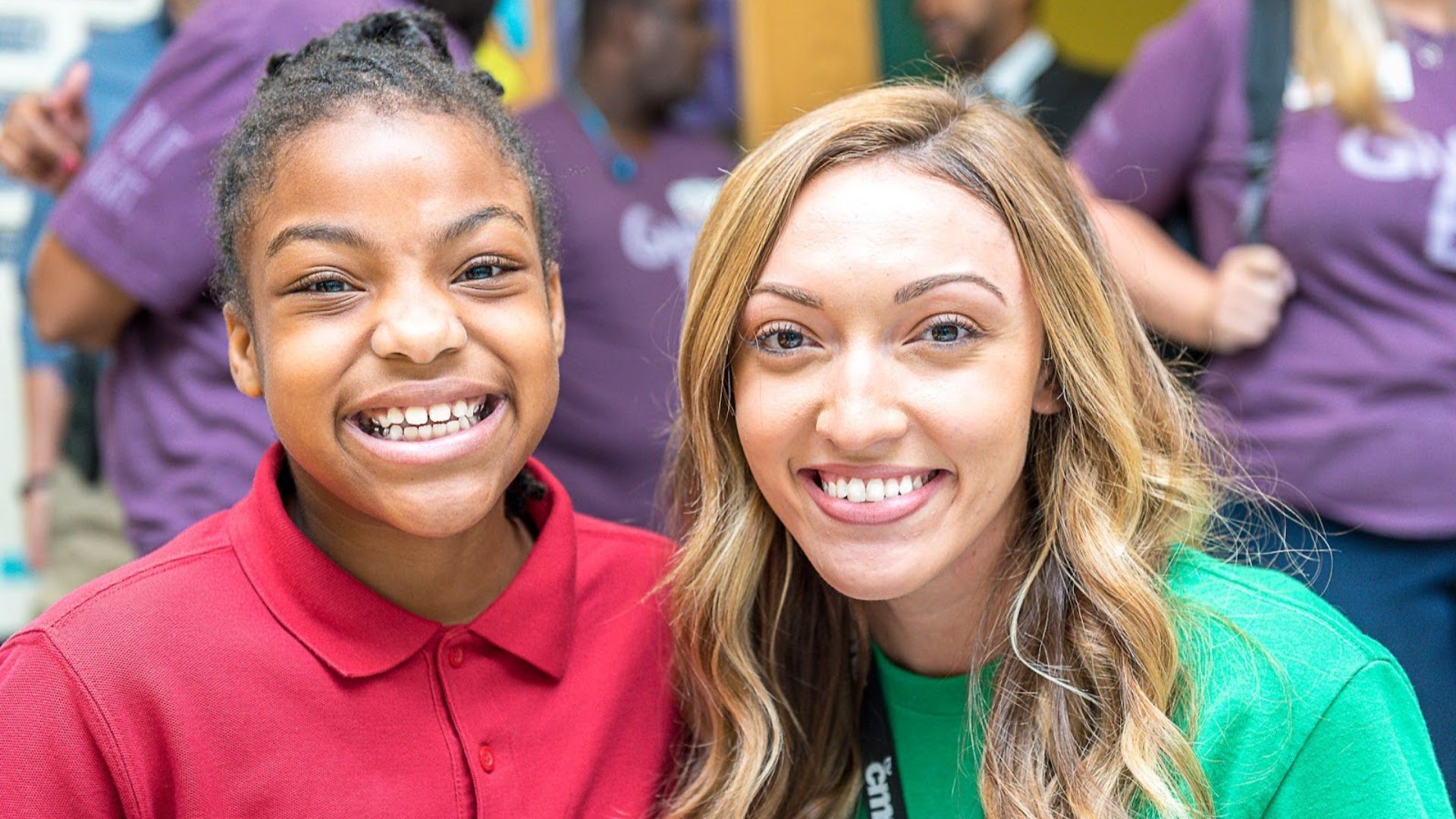 Two smiling people pose for a photo: a young girl in a red polo on the left and a woman with long blond hair in a green shirt on the right, with a blurred purple-clad background.
