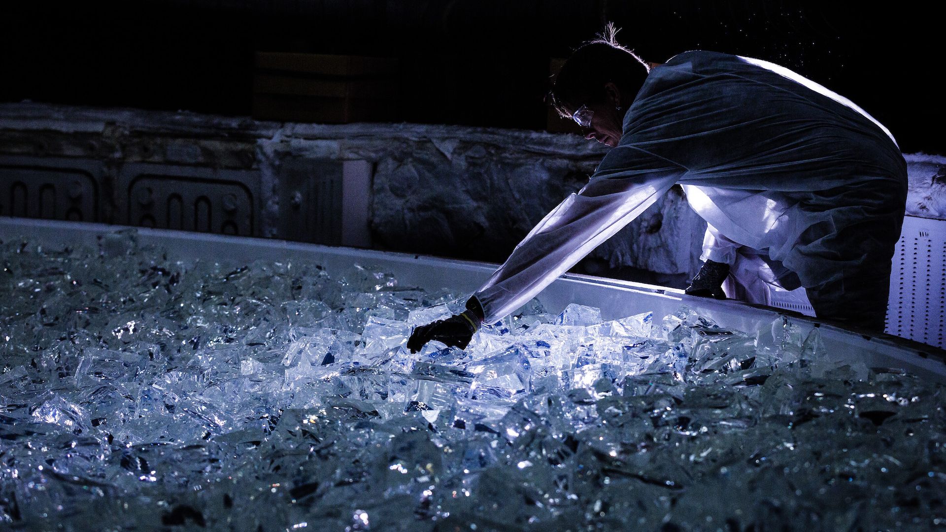 A huge plate of dimly lit glass being placed into a furnace by a man in a white clean room suit