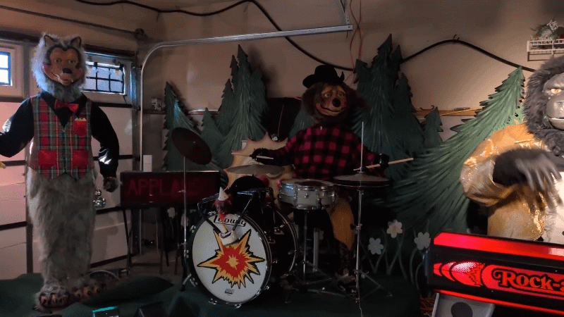 Animated GIF shows three vintage animal animatronics performing in the Howell family's Hermitage garage. A bear in a red plaid shirt plays the drums while two other characters move their arms and heads against a backdrop of artificial pine trees.