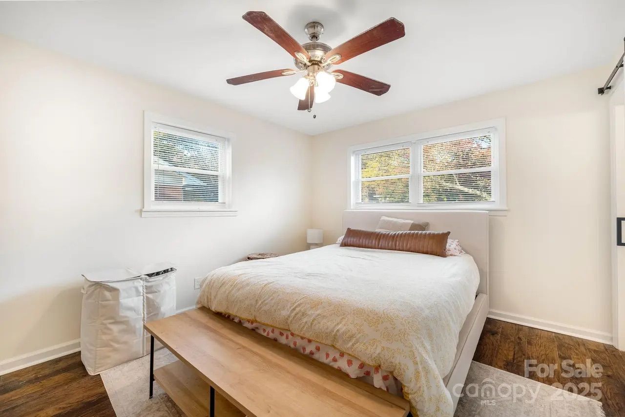 Bright bedroom with white walls, large bed with beige and brown pillows, wooden bench at foot of bed, ceiling fan with lights, two windows with blinds, hardwood floor, and laundry bins.