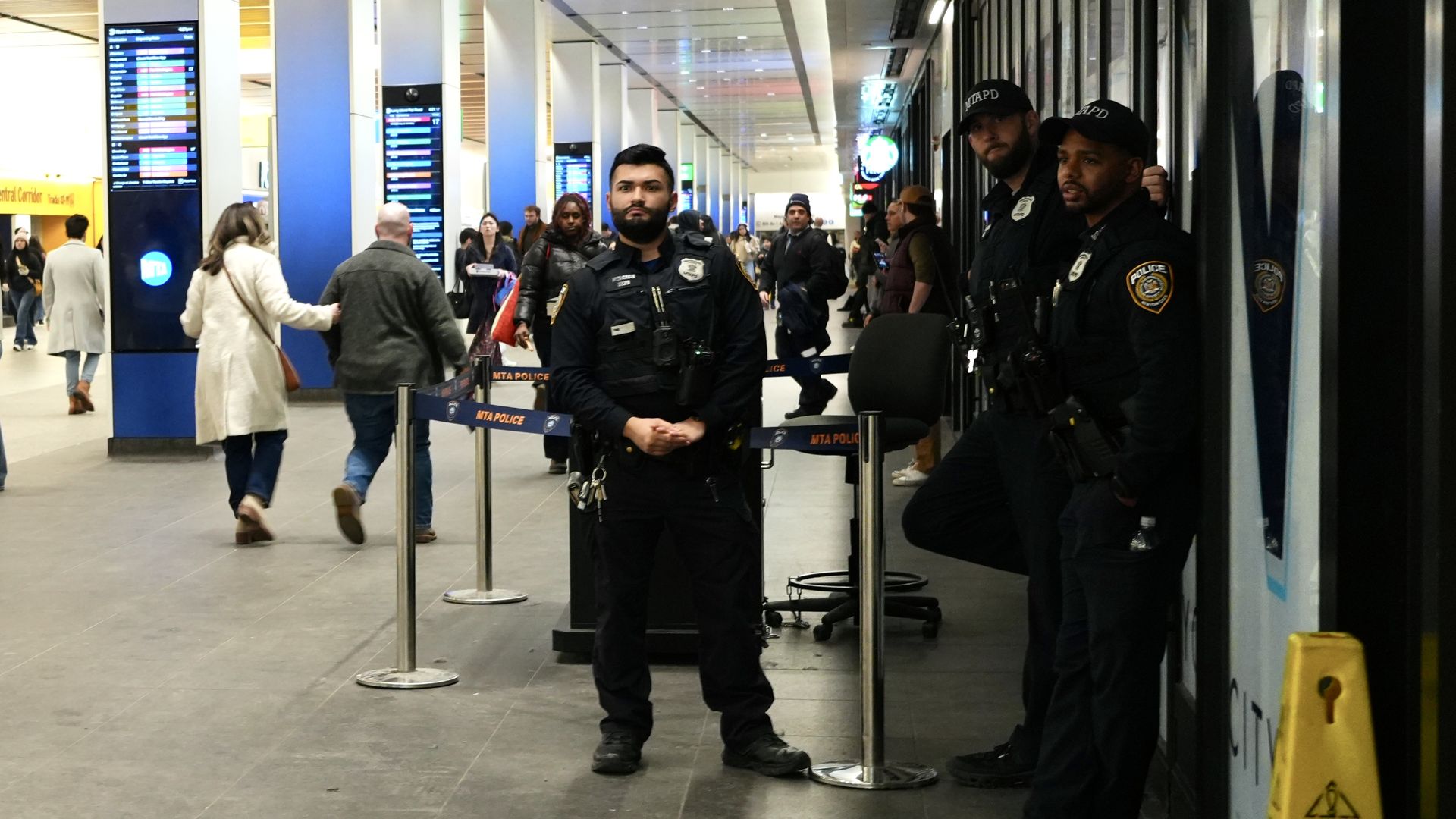 Three MTA police officers in uniform standing and leaning in a busy train station corridor with commuters walking by and digital schedule boards in the background.