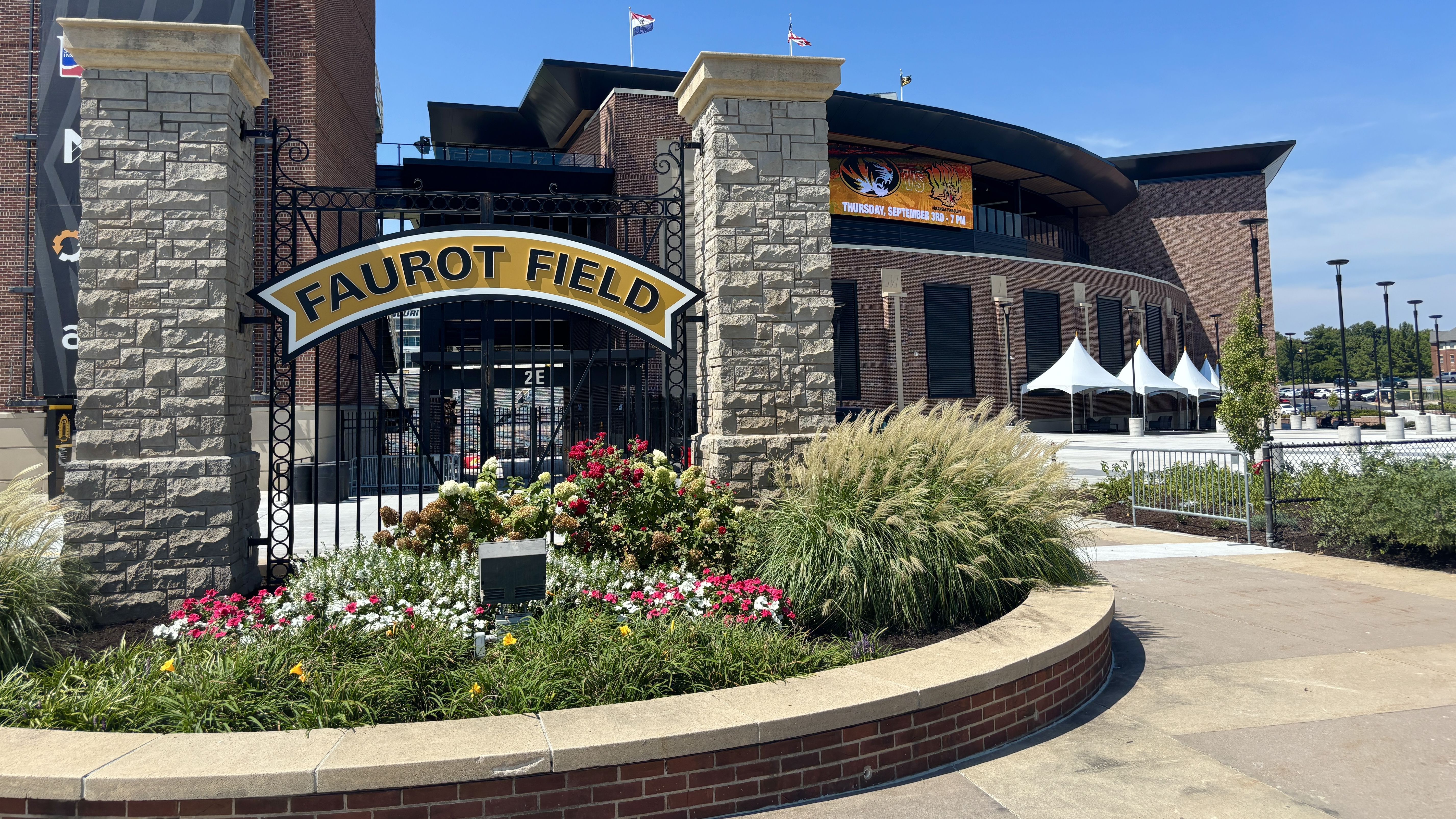 Sunny Faurot Field entrance with stone pillars, black iron gate, and a curved yellow sign reading Faurot Field. Brick building, banners, white tents, and a colorful flower bed in the foreground.