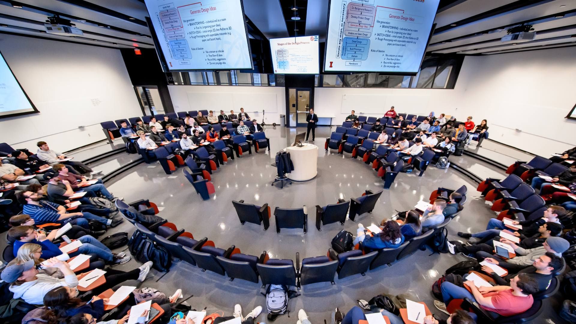 Modern lecture hall with circular, tiered seating around a central podium. Students sit in blue seats, many taking notes as large screens display slides overhead.