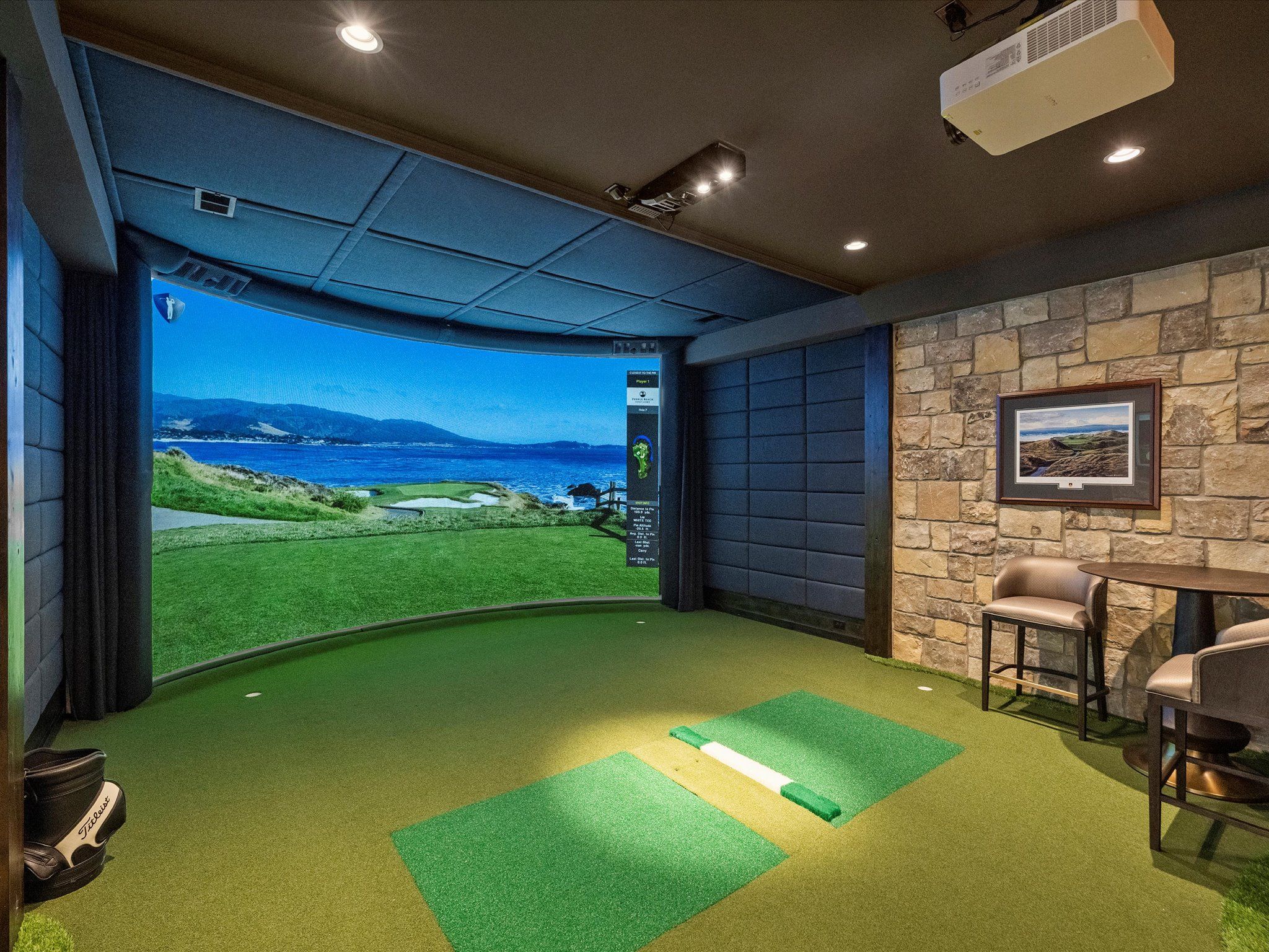 Indoor golf simulator room with a large curved screen showing a coastal golf course, green putting mats, stone wall with a framed picture, two chairs, and a small round table.