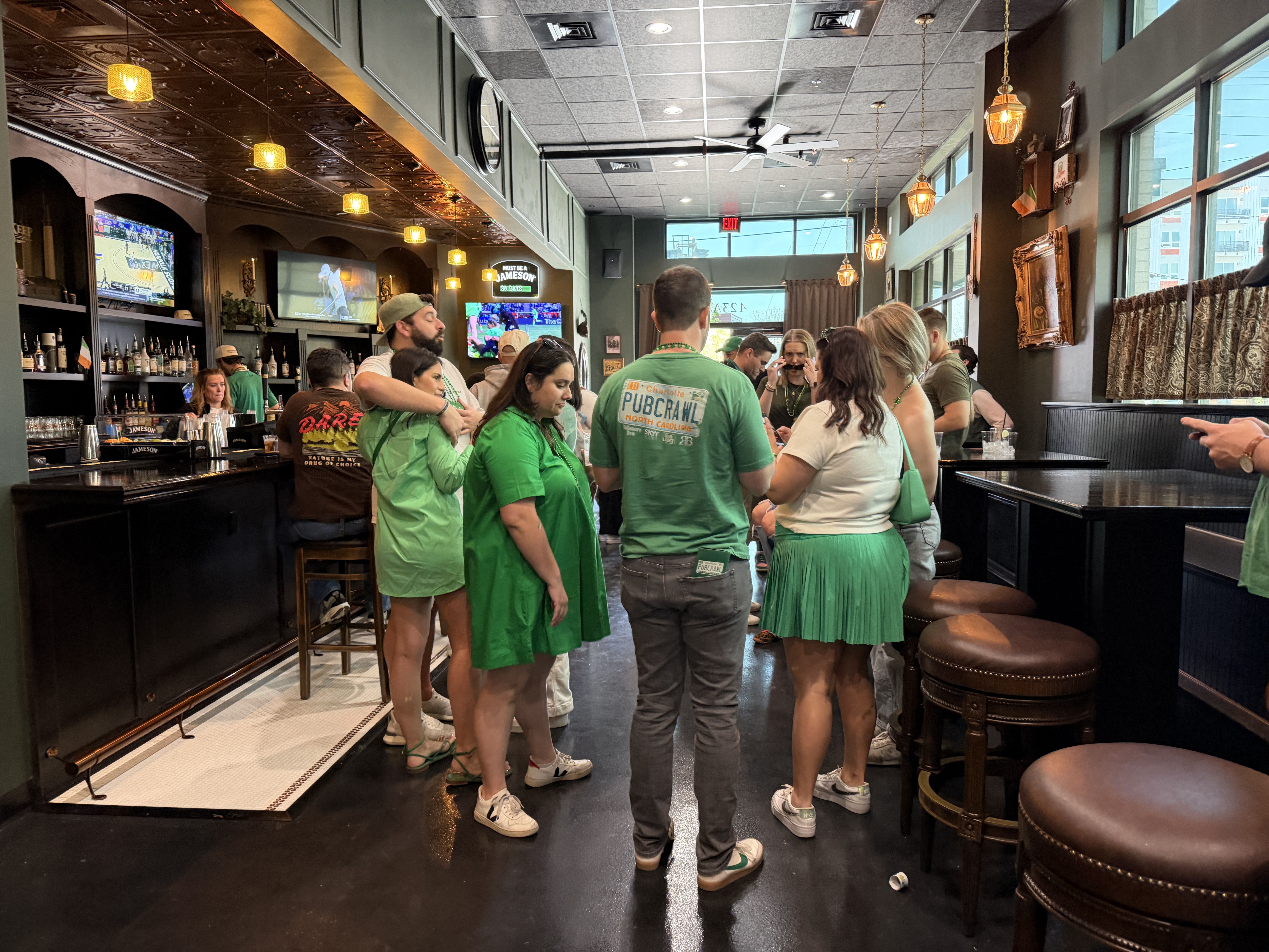 Inside a bustling bar, people in green chat near the black bar. Bottles line the left, TVs show sports, warm pendant lights glow, and large windows brighten the right side.