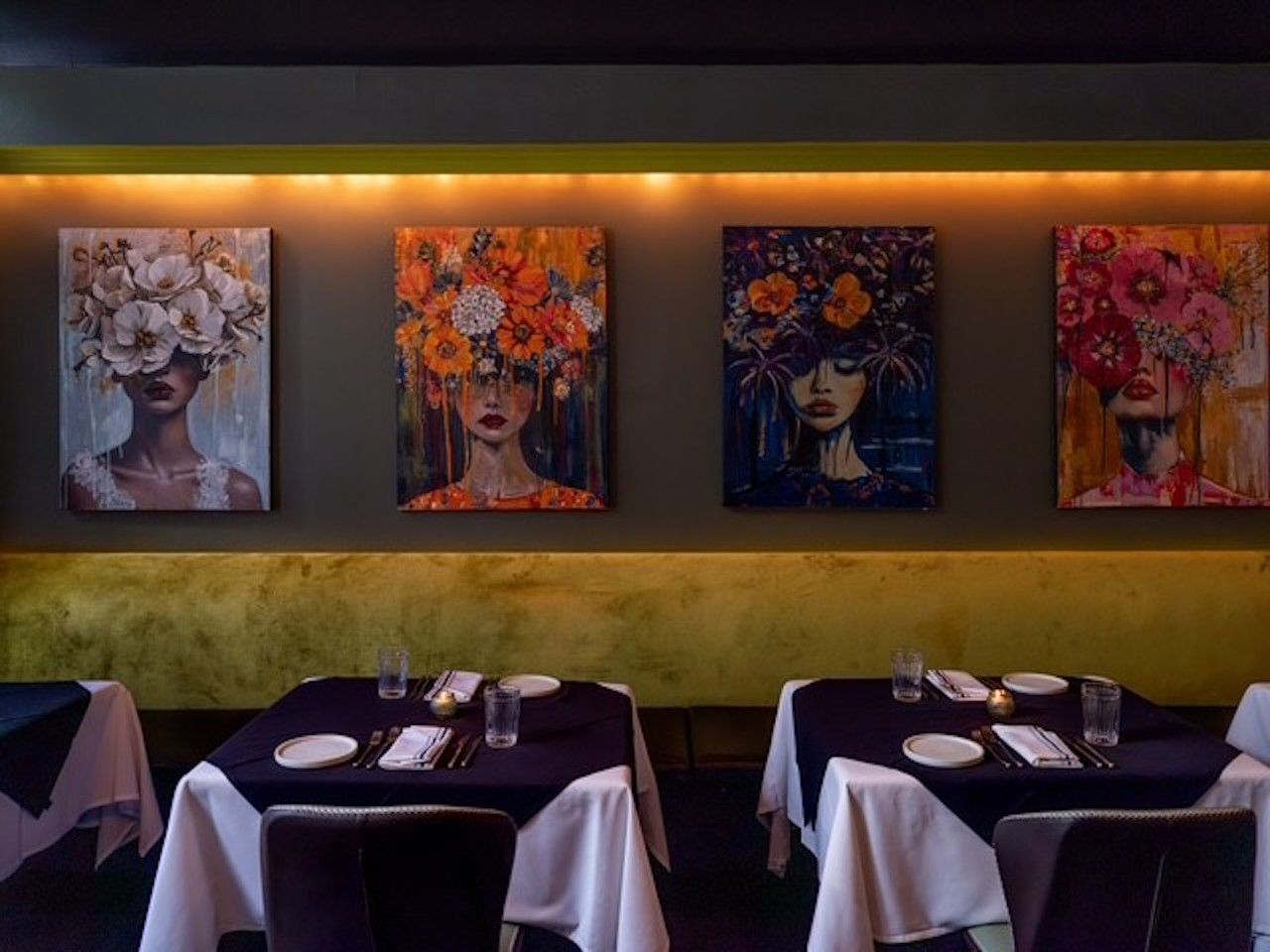 Dimly lit restaurant with four vibrant paintings of women wearing flower headdresses on a brown wall. Tables with white cloths, black runners, plates and glasses.