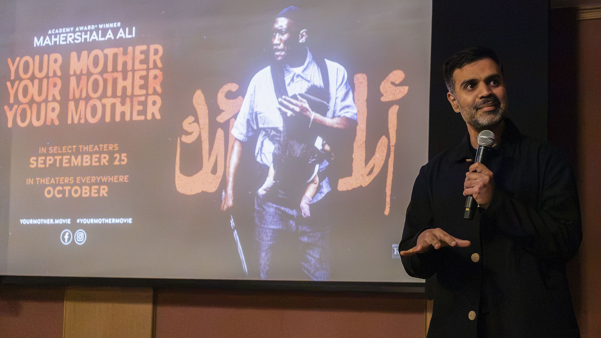 Speaker in dark clothing with a microphone stands beside a projected poster featuring orange 'YOUR MOTHER' text and a large image of a man holding a book.
