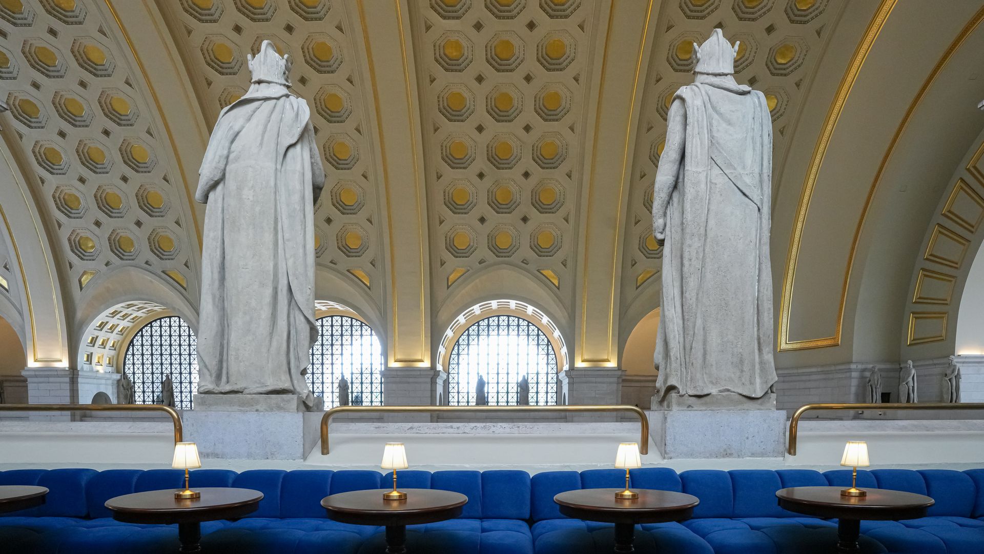 Interior of a grand hall with two large marble statues in cloaks facing away, a gold-trimmed coffered ceiling, arched windows, and blue curved seating with small round tables in the foreground.