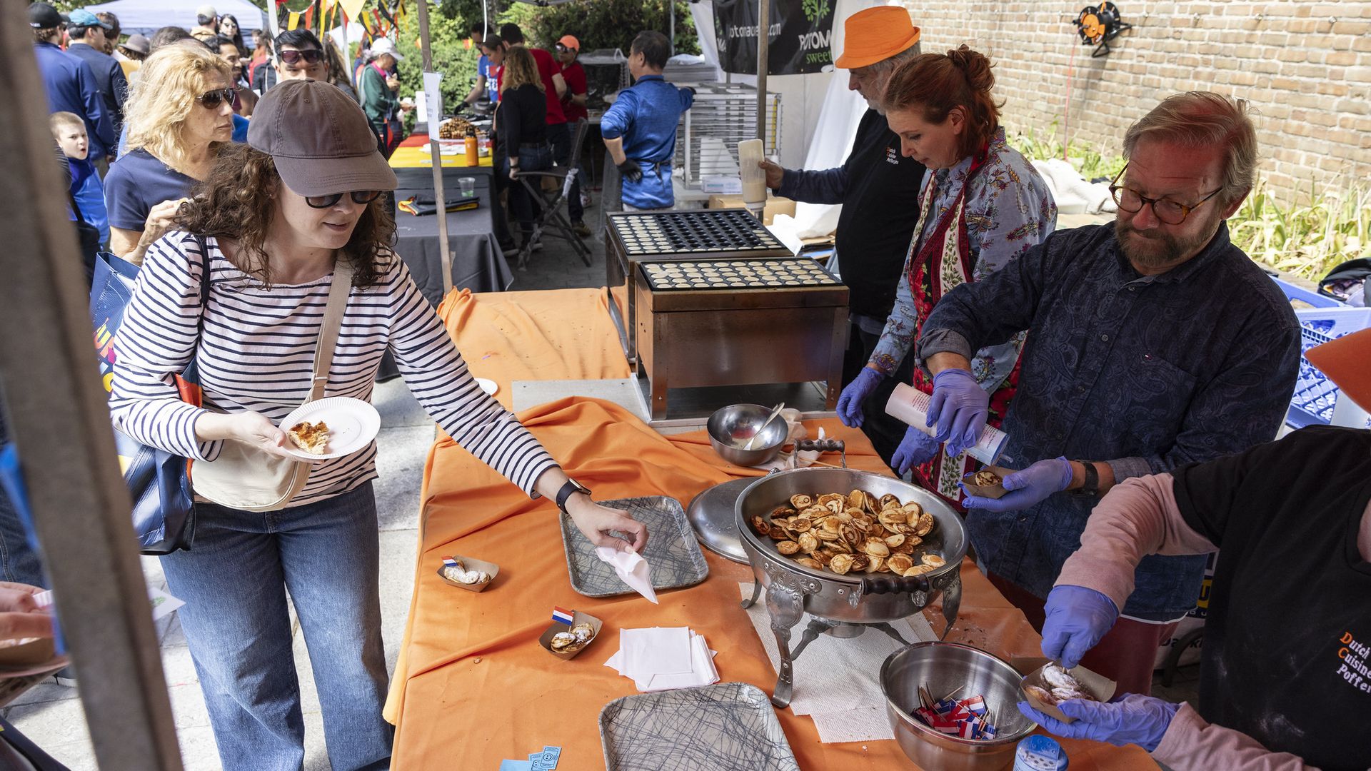 Outdoor market stall under tents with an orange tablecloth; a cook in gloves grills small round pastries as a striped-shirt shopper selects a sample, with a crowd and brick wall in the background.