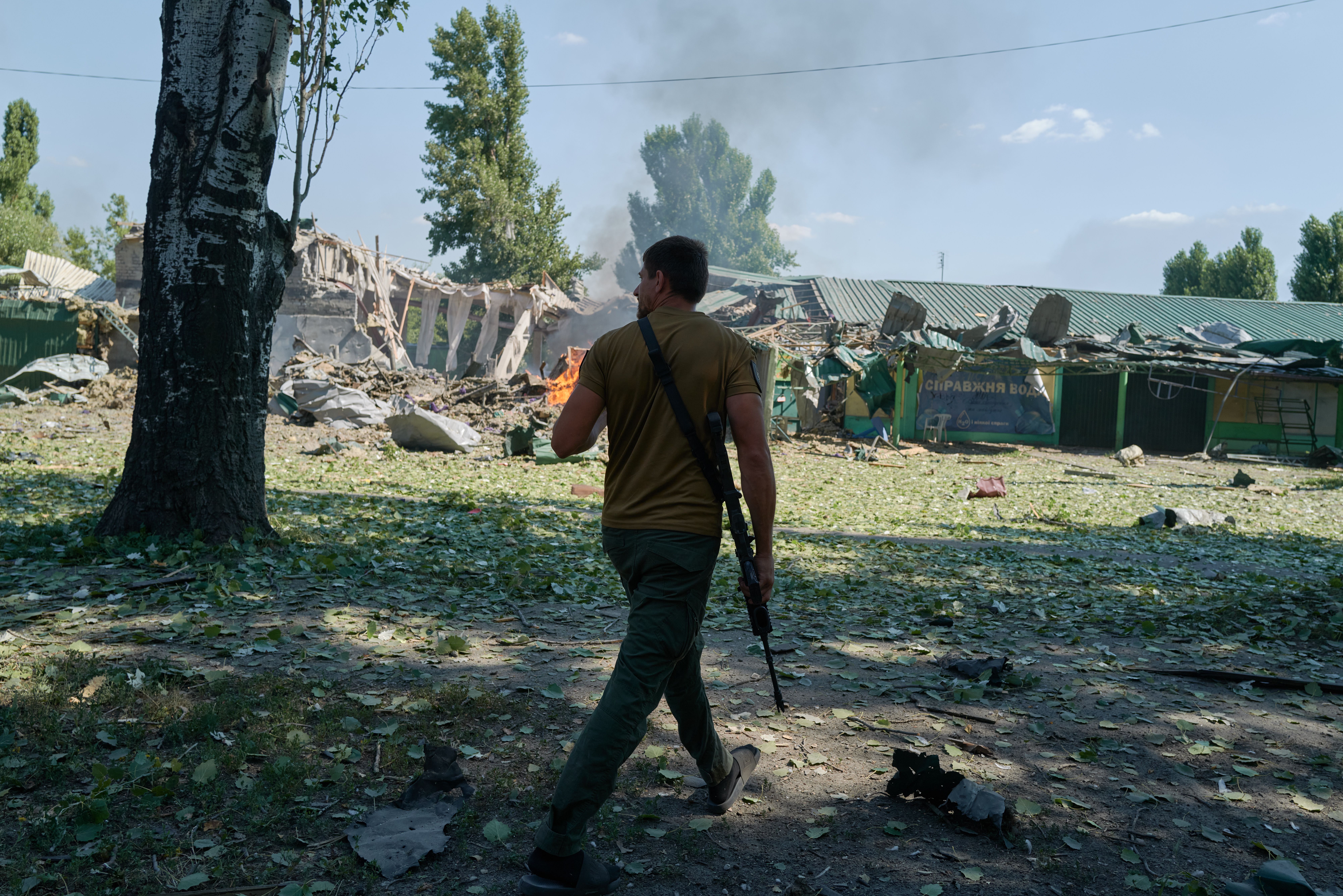 A man in a brown shirt and green pants carries a rifle, walking past debris and a fire near a damaged green-roofed building under a blue sky. Leaves and rubble cover the ground.