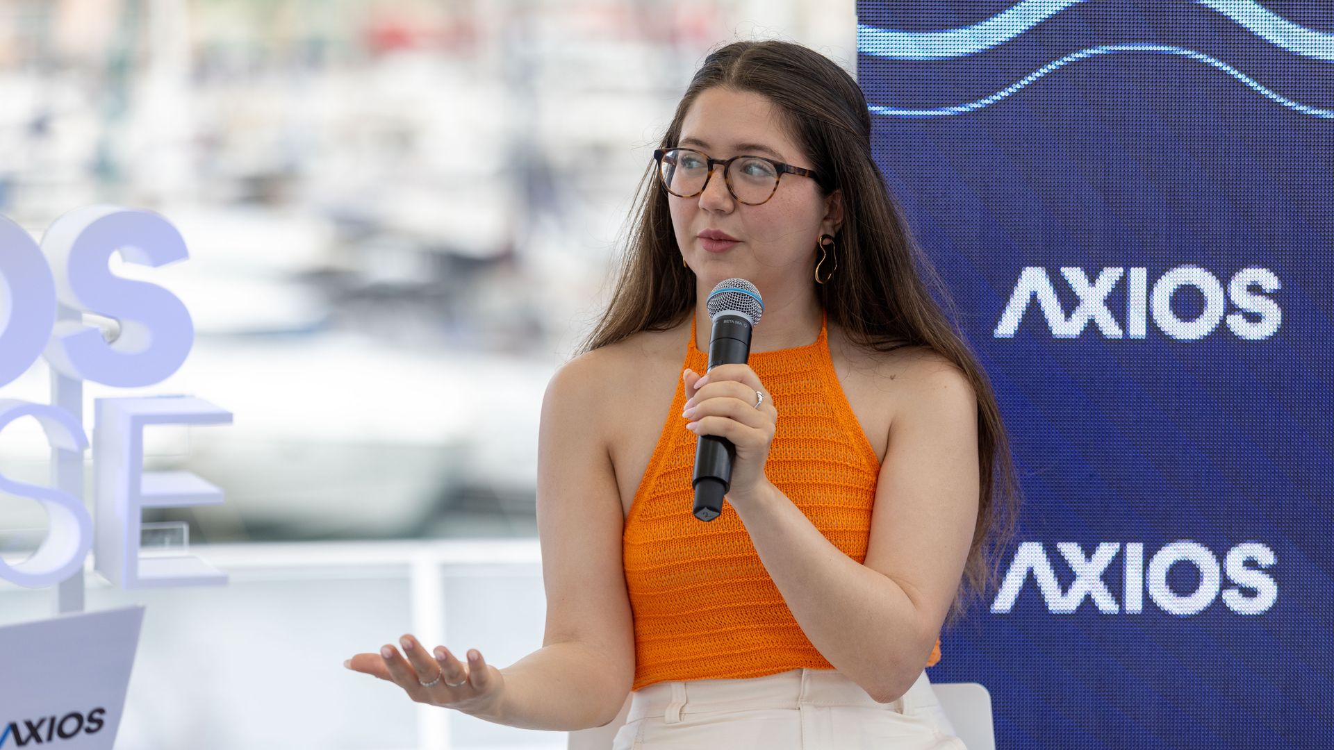 Young woman with tortoiseshell glasses and an orange knit top speaks into a handheld microphone on a stage, beside a blue AXIOS backdrop.