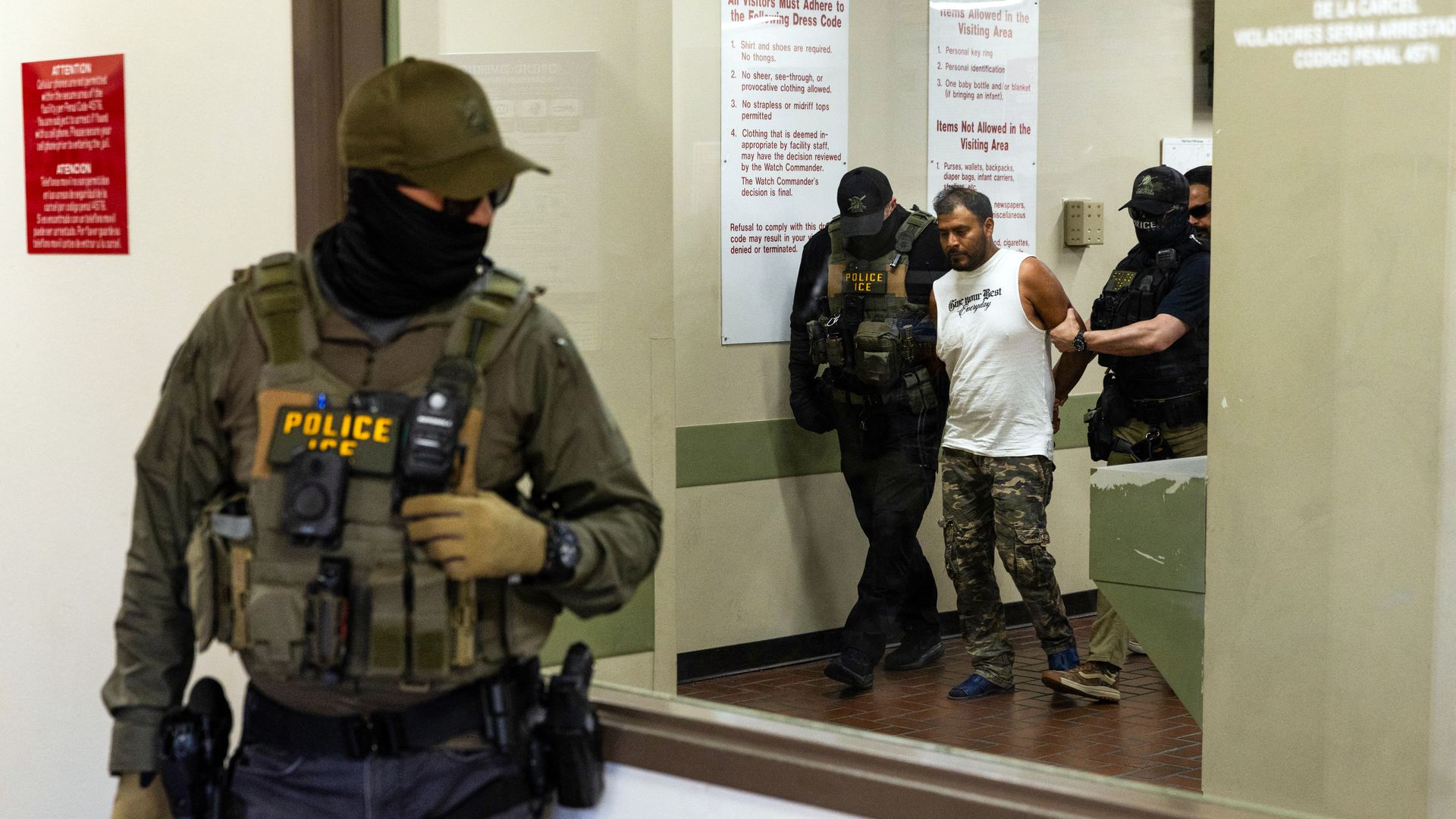 A man in a white tank top and camo pants is walked out of a building by ICE agents while his hands are behind his back. Another ICE agent, whose face is covered, stands in the foreground.