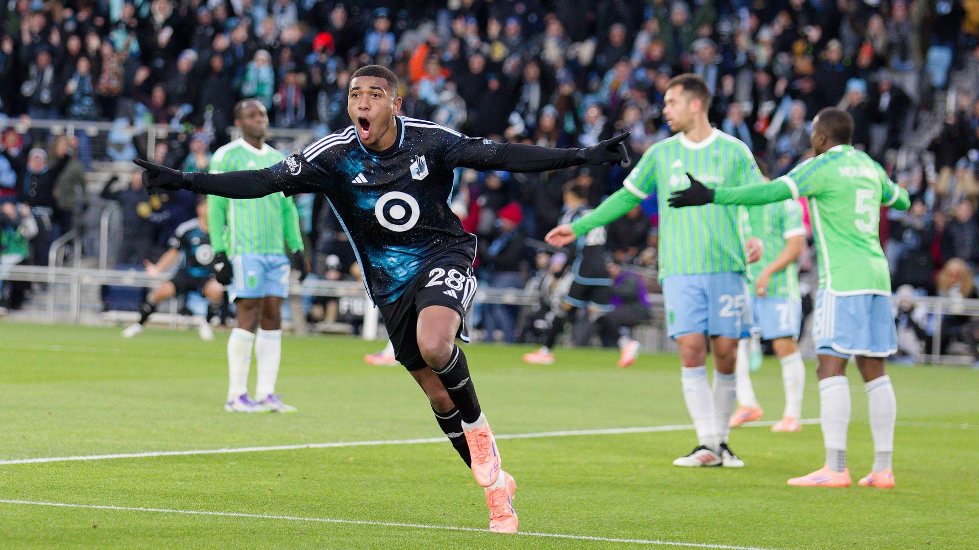 A soccer player in a black uniform with a target logo celebrates with his hands out as three opposing players in green jerseys look at each other in confusion. 