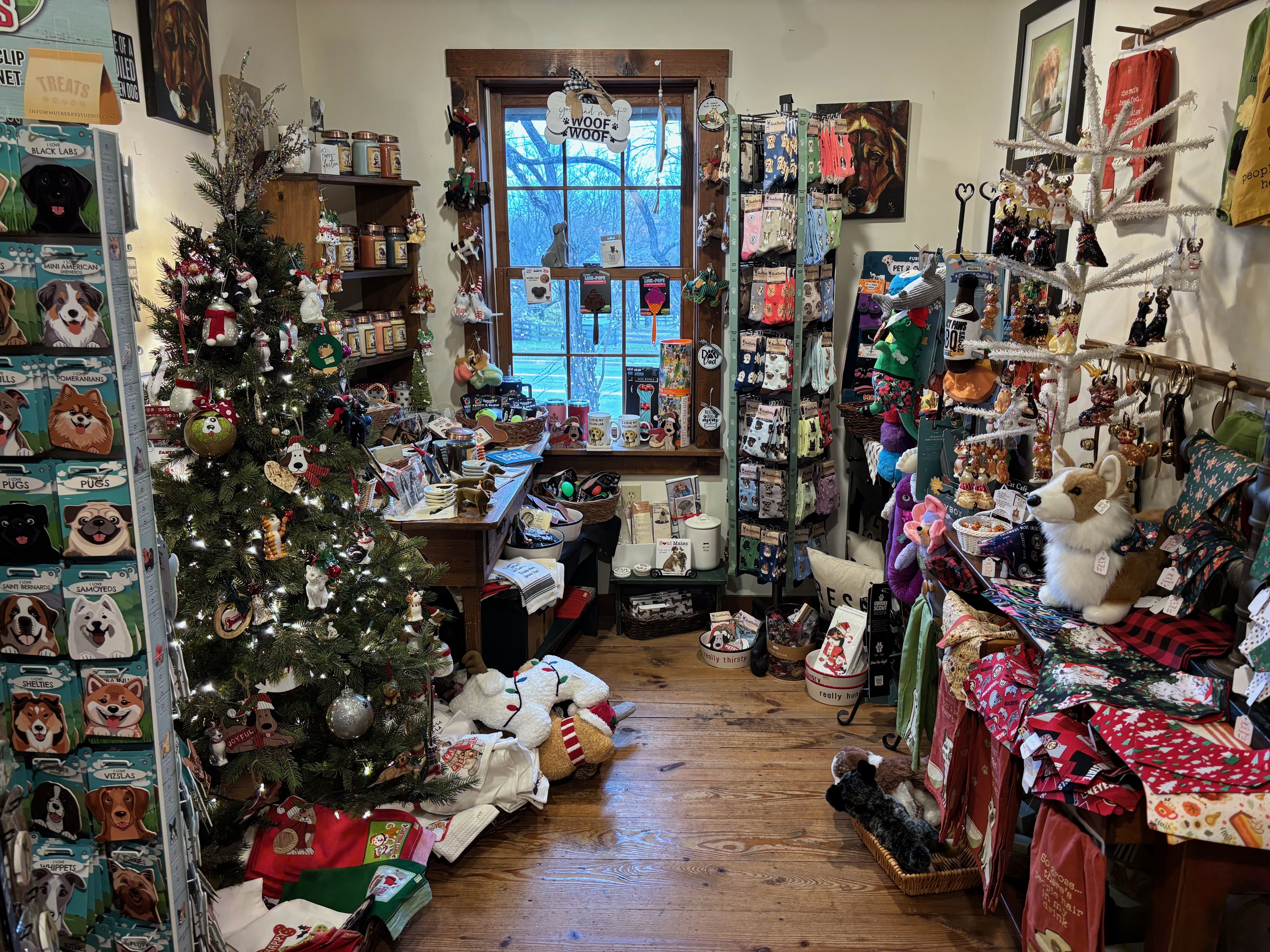 Cozy gift shop corner decorated for Christmas with a lit tree, various cat and dog-themed items, socks, ornaments, mugs, and stuffed animals displayed on shelves and racks around a window.