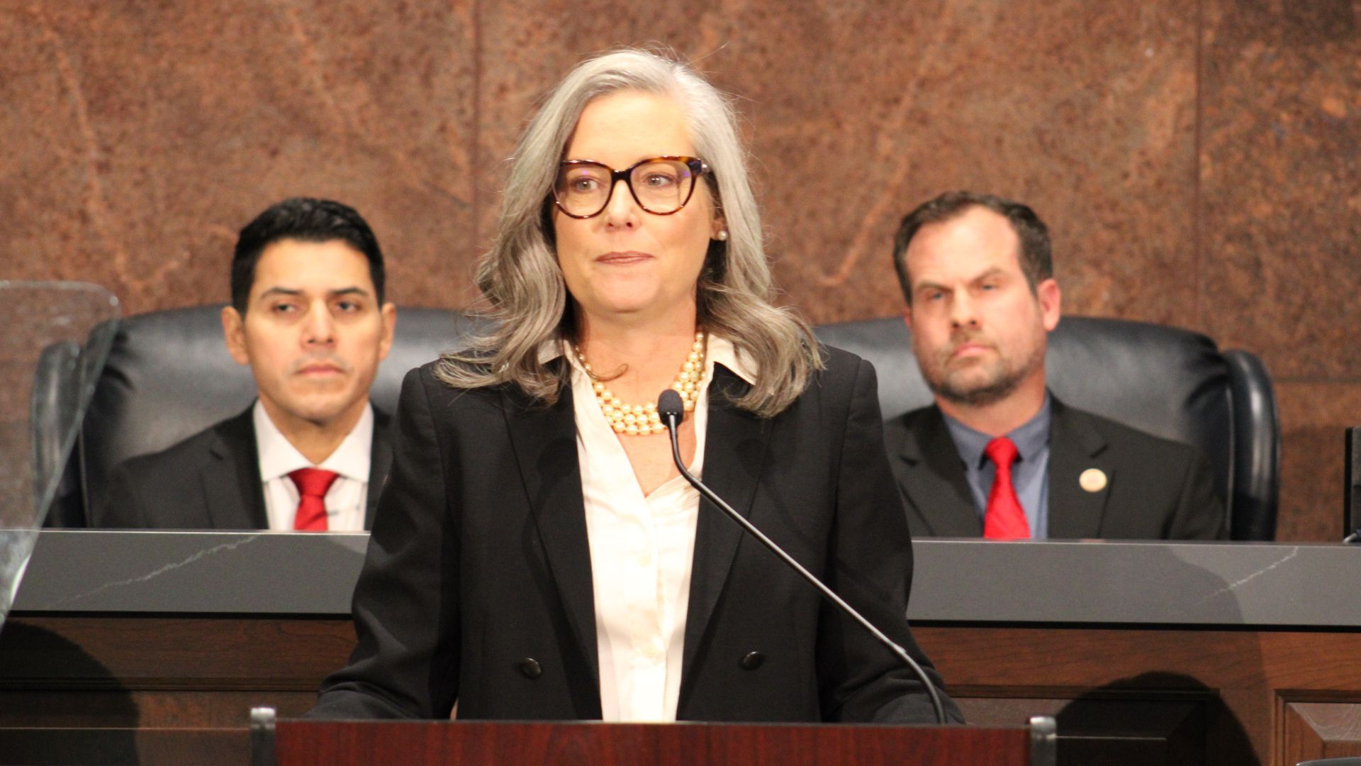 Katie Hobbs stands on a dais in front of a microphone, with Steve Montenegro and Warren Petersen seated behind her.