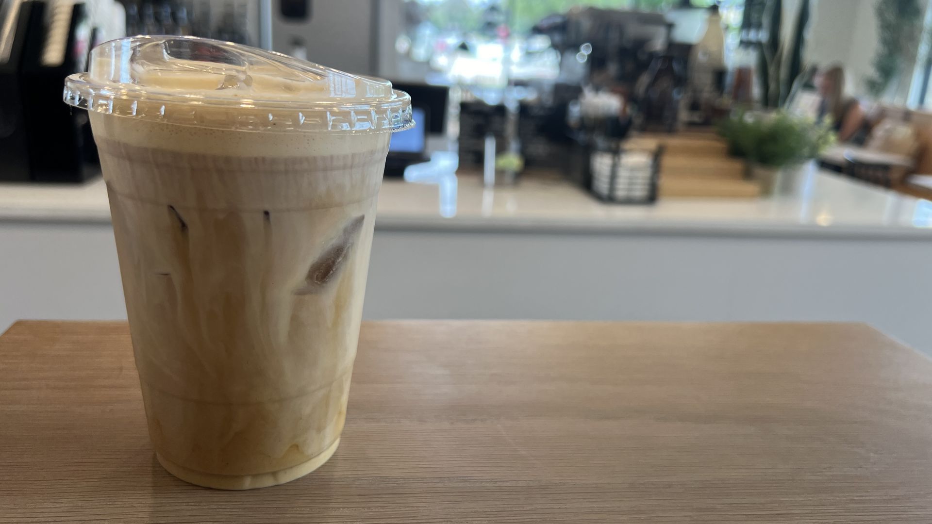 Iced coffee with cream swirls in a clear plastic cup with lid on a wooden table inside a bright cafe.