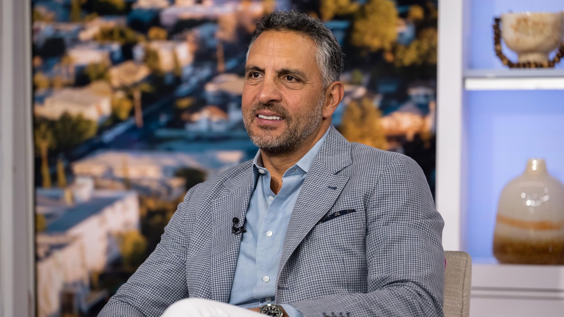 A photo showing Mauricio Umansky sitting in what looks like a homey studio set wearing an open blazer and buttoned down shirt with the top two buttons undone. He's slightly smiling. He has a stubble beard and salt and pepper hair.