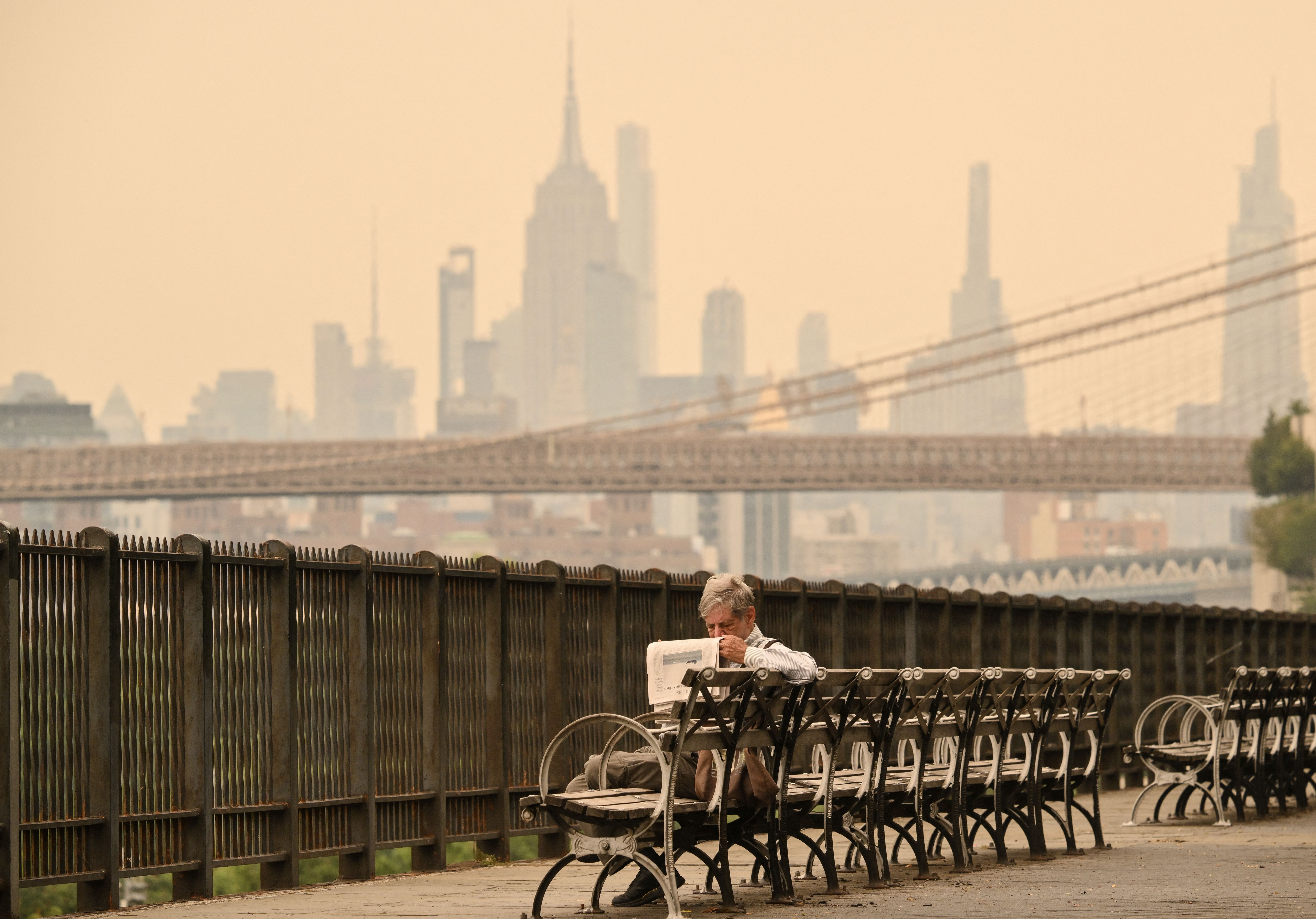 A person on a bench reads a newspaper and the hazy skyline of Manhattan is visible in the background