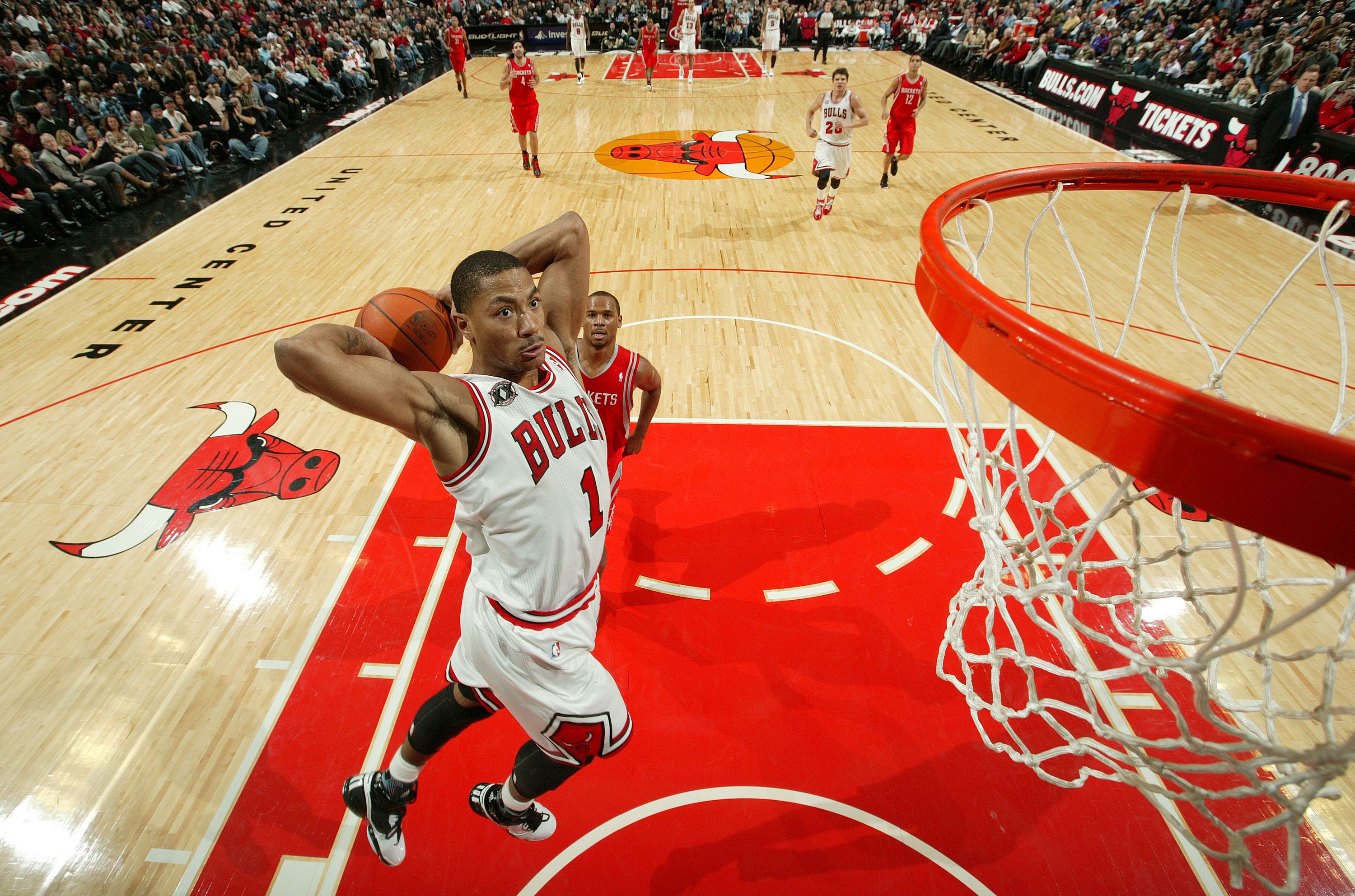 Chicago Bulls player in white uniform leaps near basketball hoop for a dunk during a game against Houston Rockets in red uniforms on a polished court at United Center.