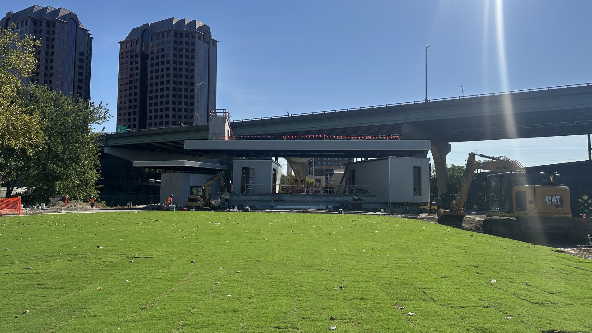 Sunny day at a construction site with two tall buildings on the left, an elevated highway across the middle, a green lawn in the foreground, and heavy machinery on the right under a blue sky.