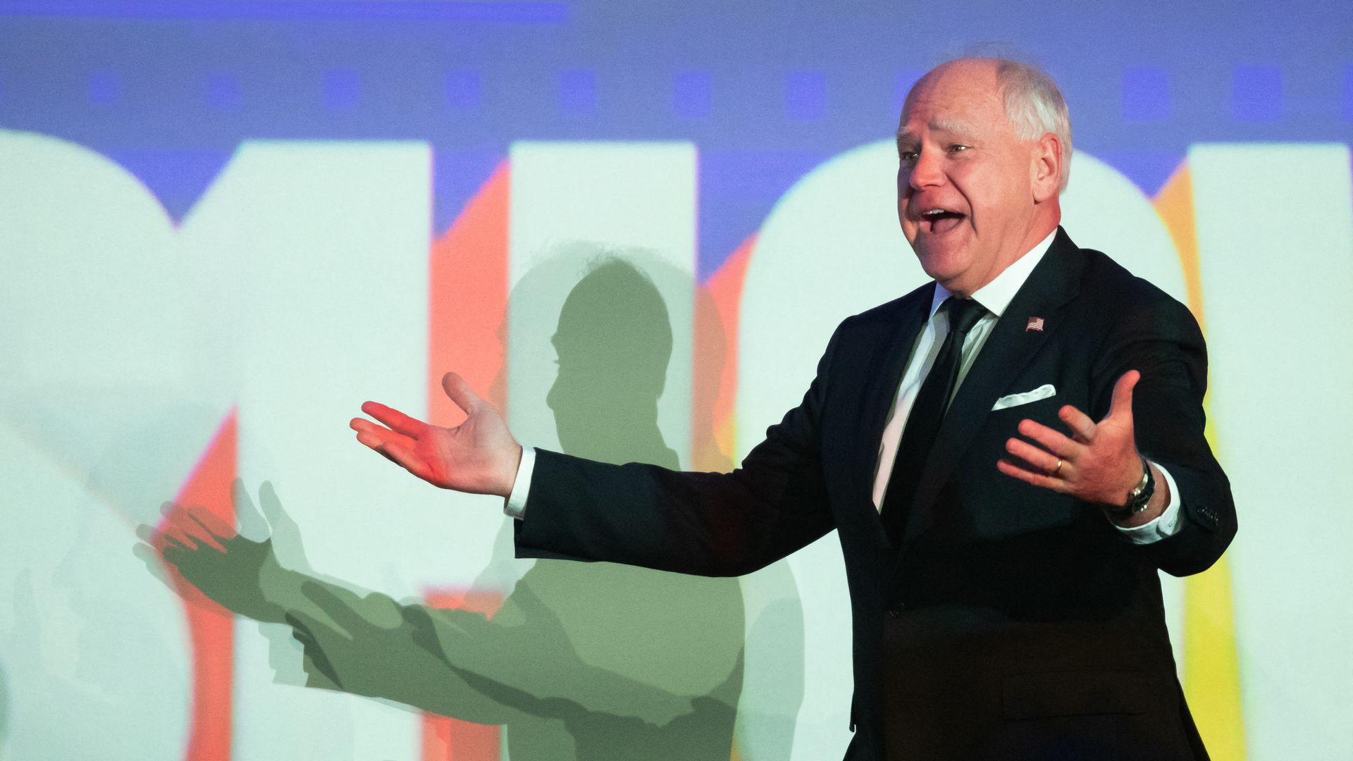 Tim Walz walks across a stage in a dark suit with his arms outstretched and an engaged expression