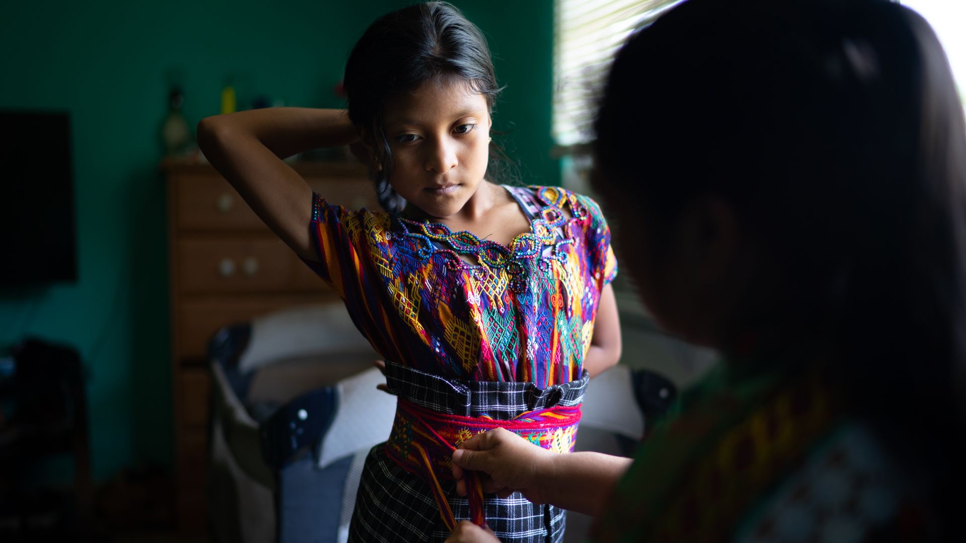 A young girl is trying on a colorful, traditional Guatemelan outfit. She has one arm up and another behind her back. two hands are trying the dress from the front but the person doing the tying is not visible. 