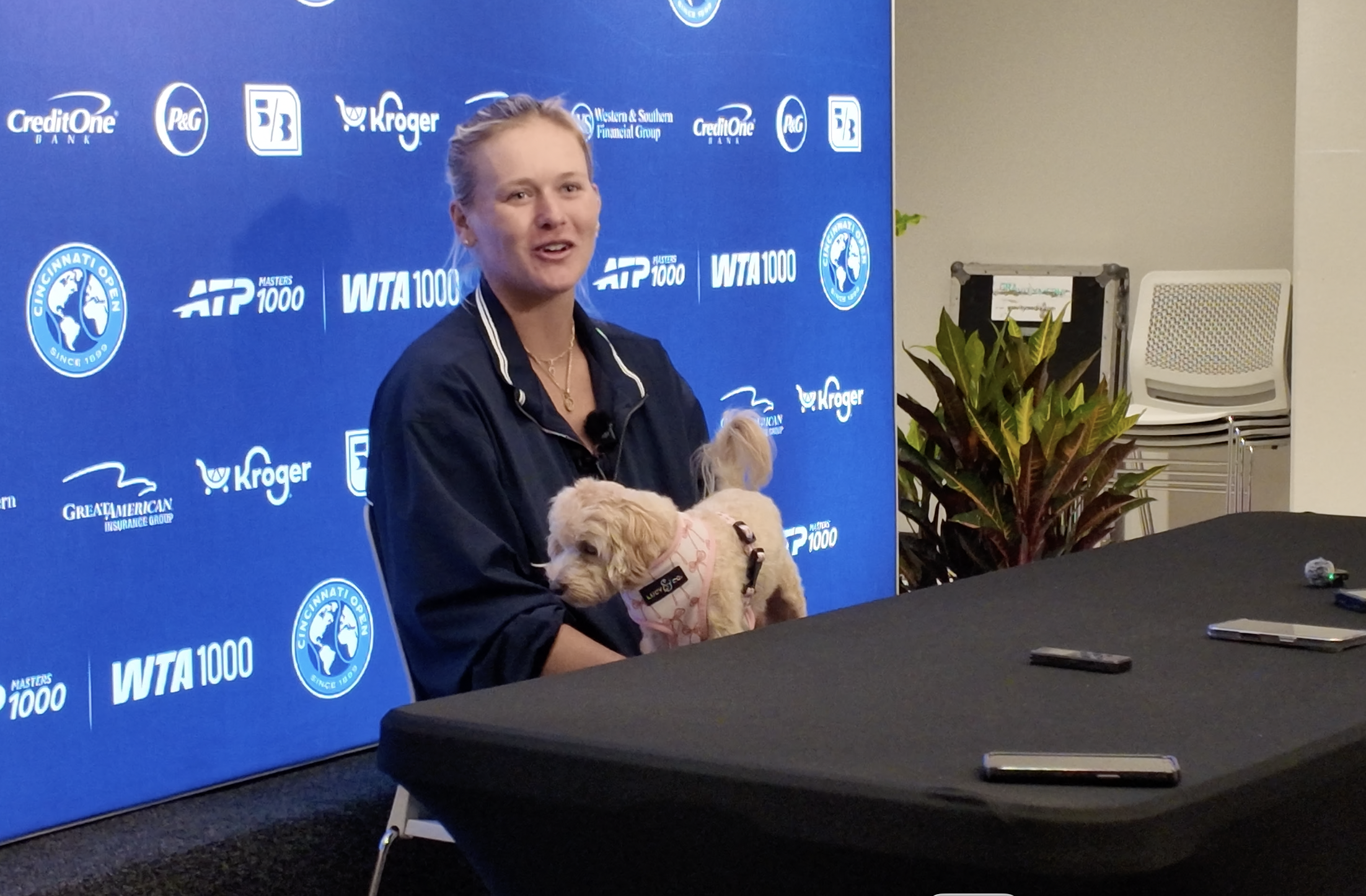 Portrait of a woman in a dark jacket seated at a table with a small white dog; a blue backdrop behind her shows sponsor logos such as ATP 1000 and WTA 1000.