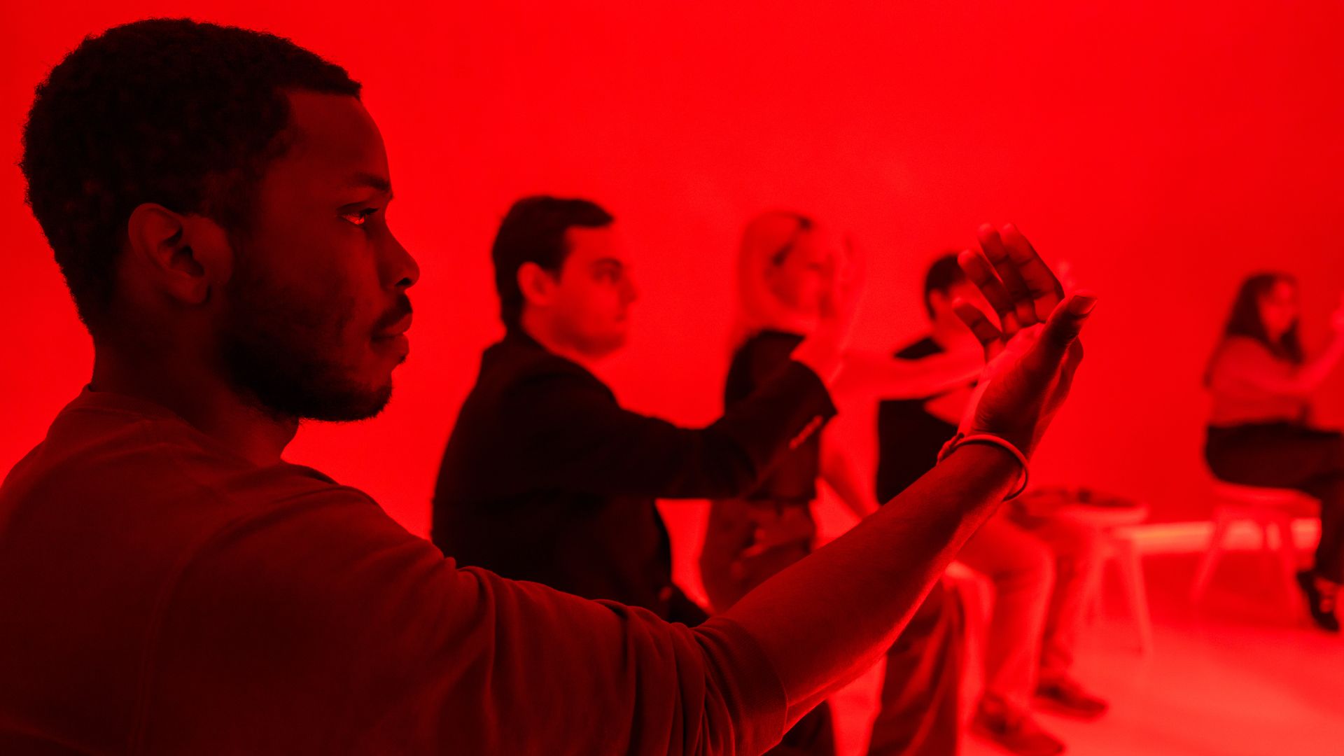 In a room bathed in red light, a man in the foreground extends his arm toward the camera. Several people sit or stand in the background, engaged in a group activity.