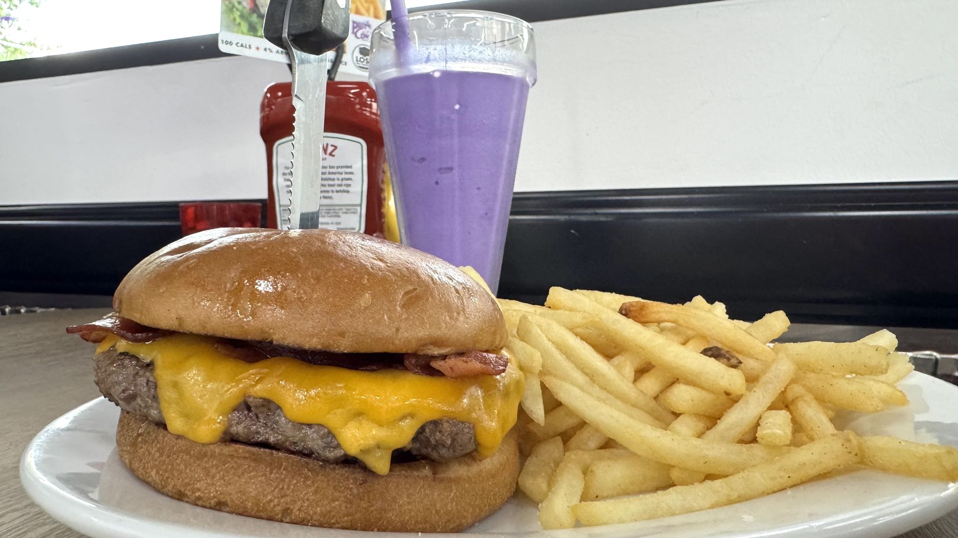 A cheeseburger with melted cheddar and bacon on a soft brown bun, with a side of fries and a tall purple milkshake with a straw in the background.