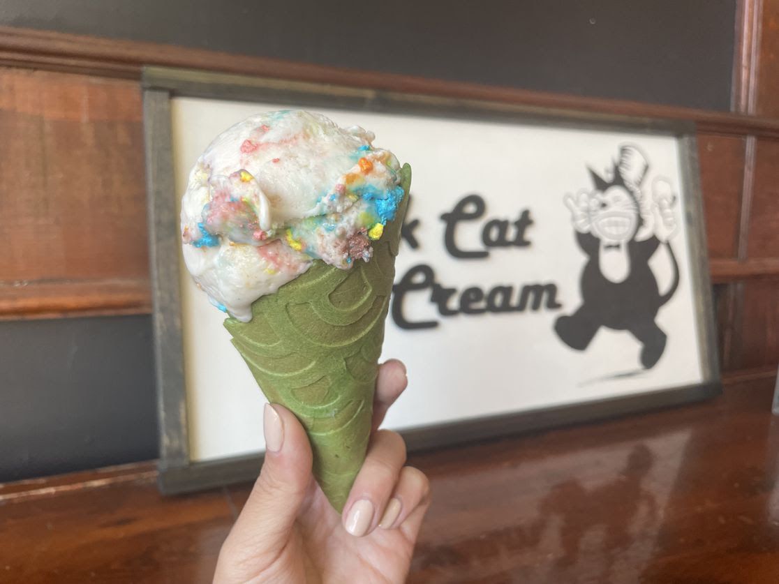Lucky Charms-infused ice cream in a green cone, held up in front of the Black Cat Ice Cream sign in Des Moines.