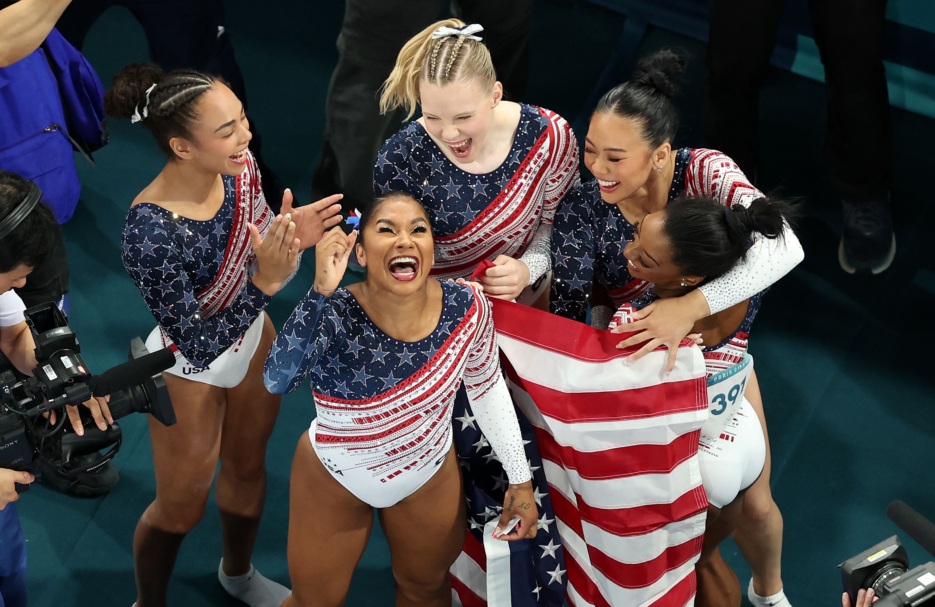 Five women in USA-flag themed leotards smile, clap and hug while one is wrapped in an American flag.