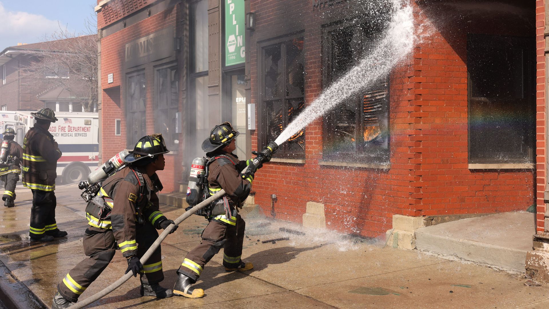 Firefighters in full turnout gear battle a blaze at a brick storefront, spraying a high arc of water into burning windows; an ambulance and other firefighters stand nearby.