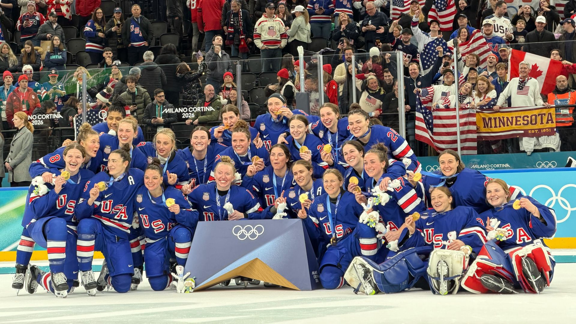 Team USA women's ice hockey players in blue jerseys celebrating with gold medals on ice rink, fans behind holding USA flags and Canadian flag, Olympic rings visible.