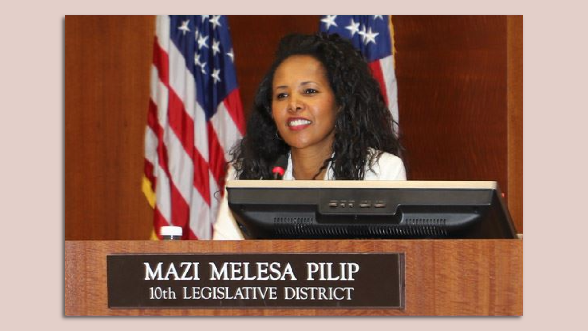 Mazi Melesa Pilip smiles behind a sign that says her name and "10th legislative district." The American flag is behind her. 