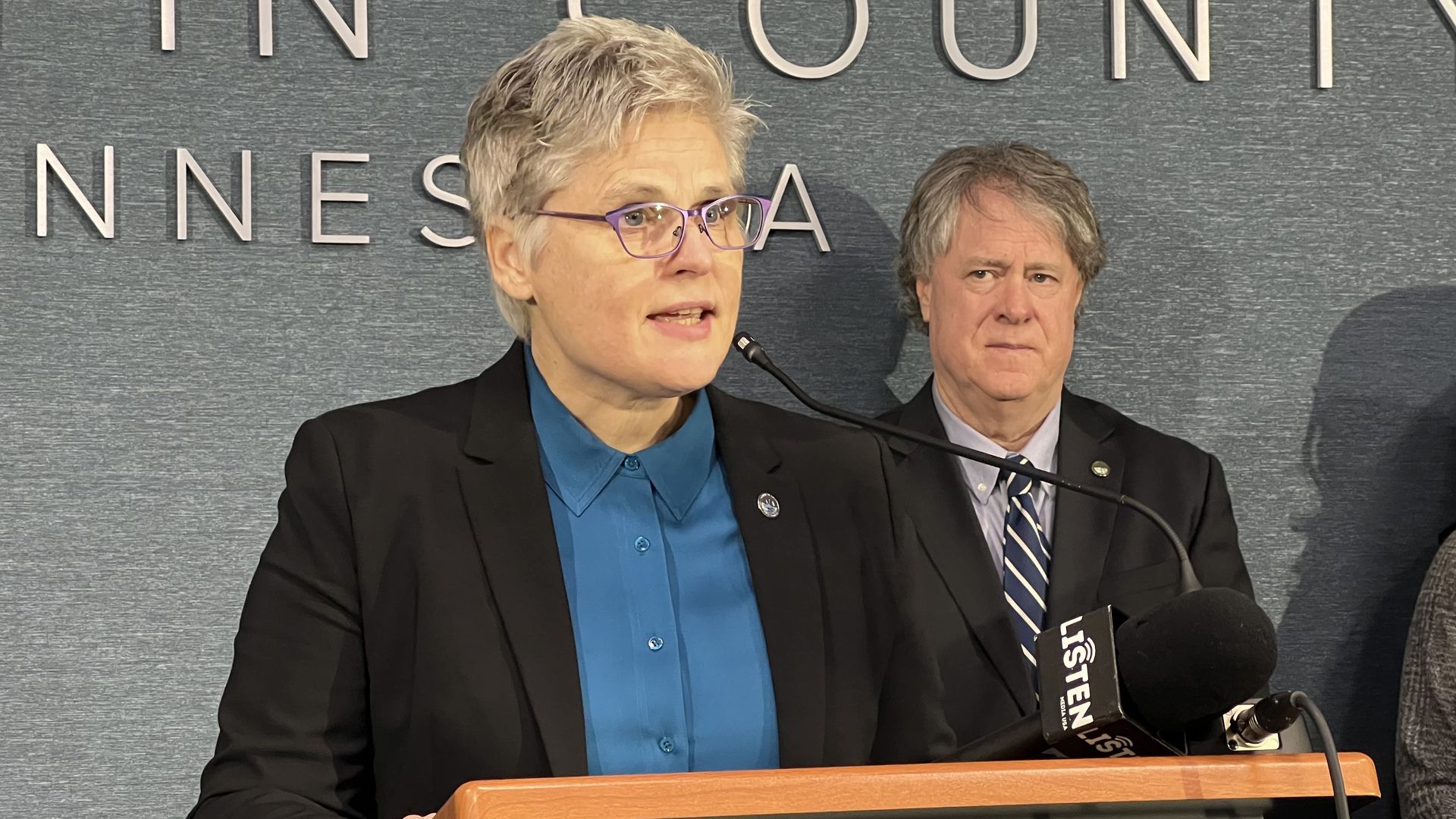 A woman with short, grey hair and glasses wearing a black suit and blue button-down speaks into a microphone as another man looks on from the background.