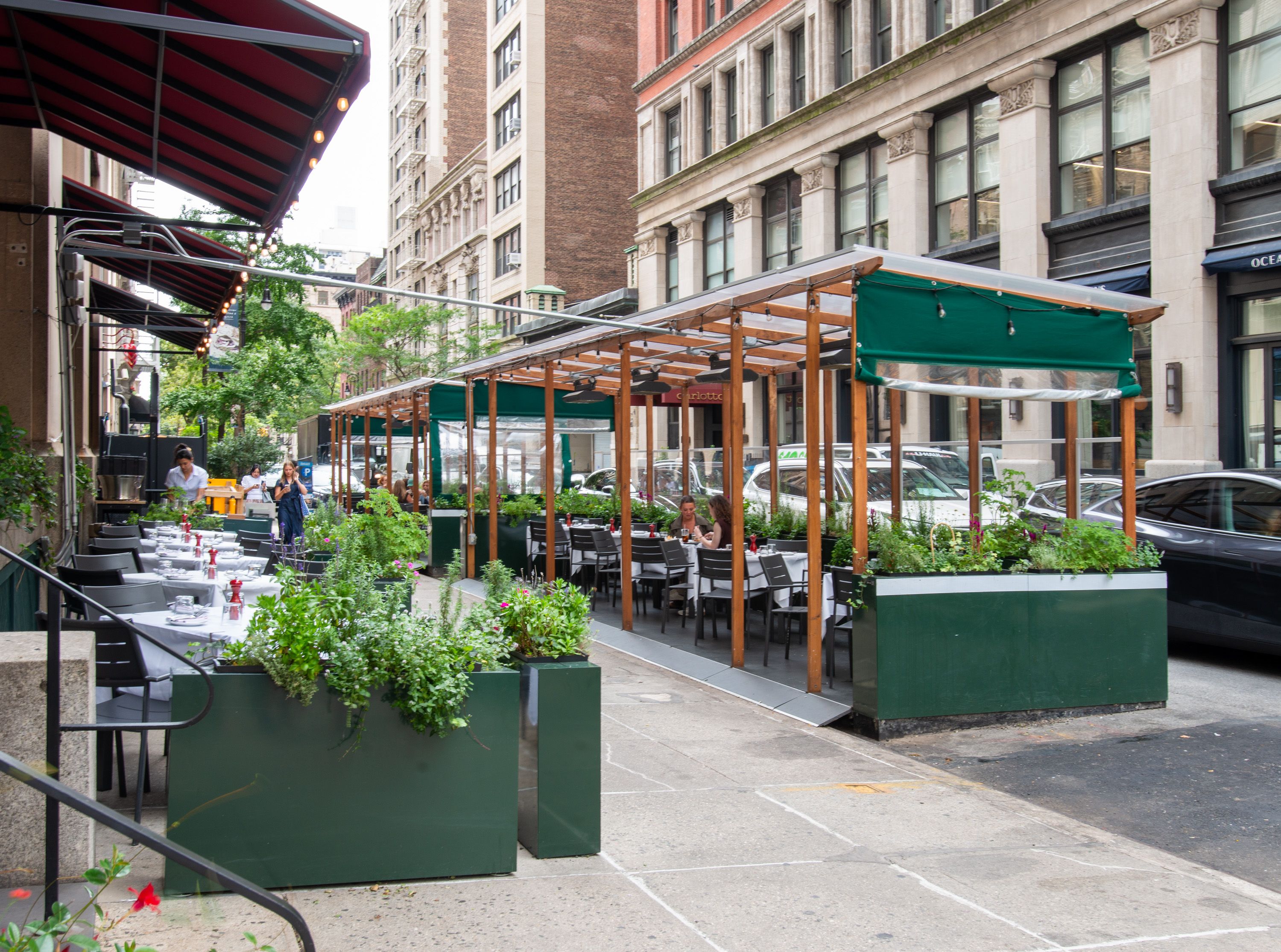 Outdoor sidewalk cafe in a city: white-clothed tables line the street under red-striped awnings; wooden-framed green planters, diners seated, tall brick buildings and parked cars in the background.