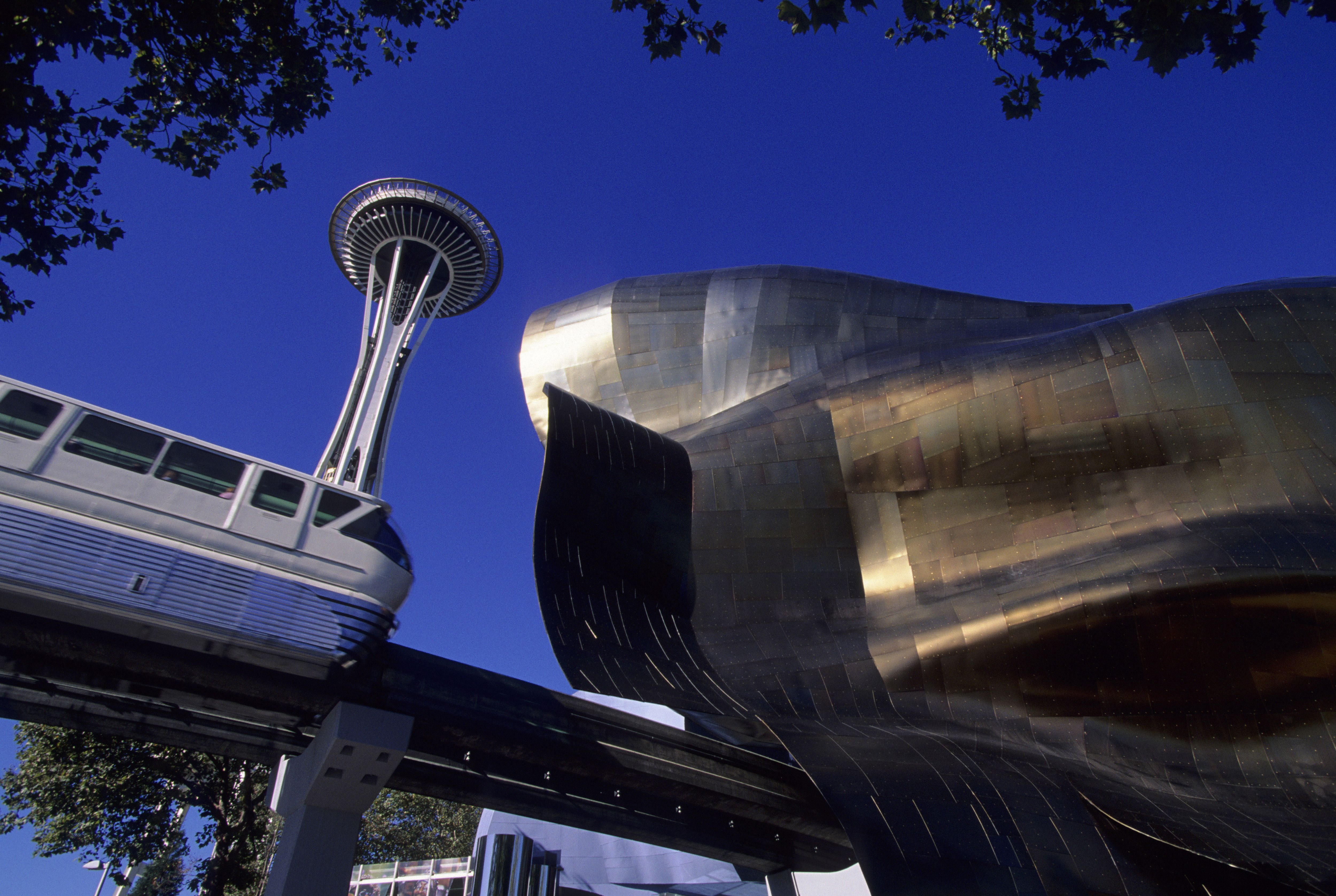 The Space Needle towers over the Seattle monorail track and part of the Museum of Pop Culture, with a blue sky behind.