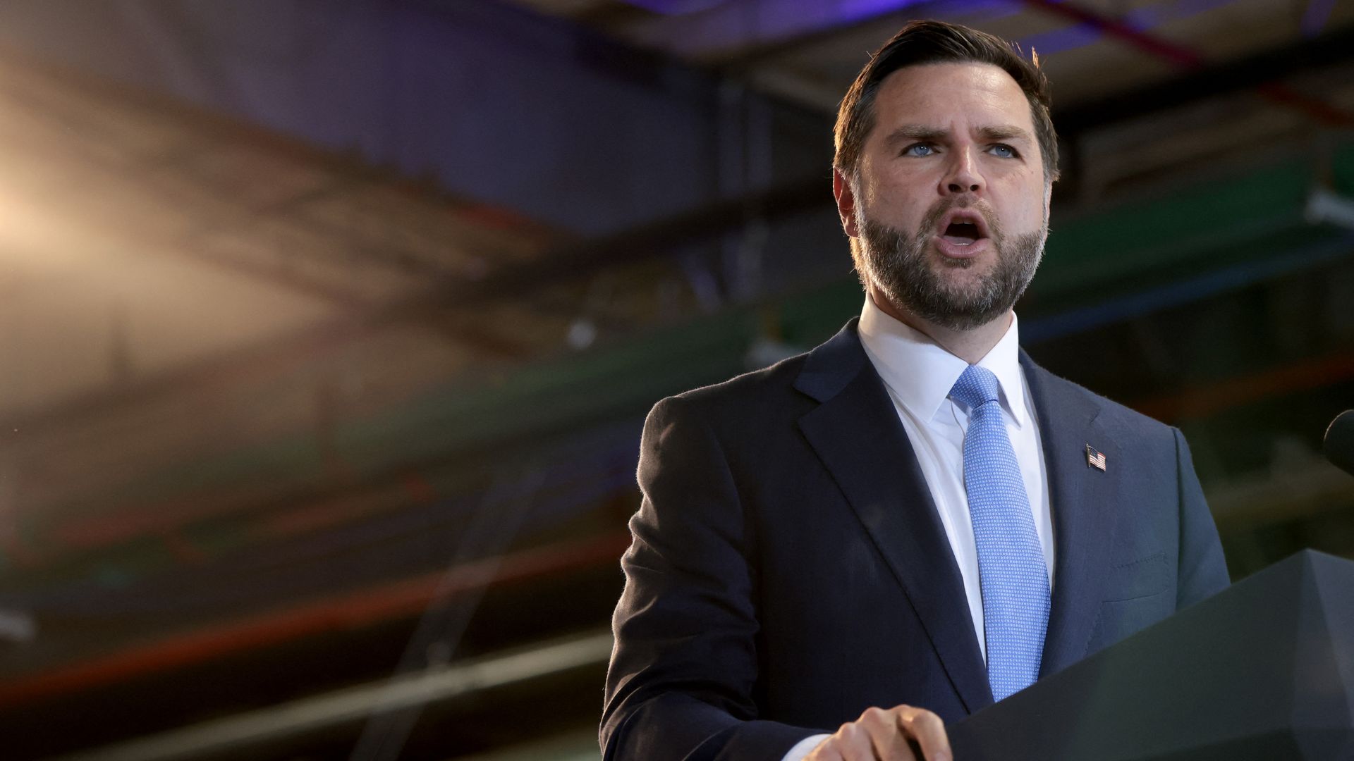 Vice President JD Vance — a man with brown hair and a brown beard wearing a navy blue suit, a white collared shirt, a light blue tie and an American flag pin — grips a podium as he delivers a speech.