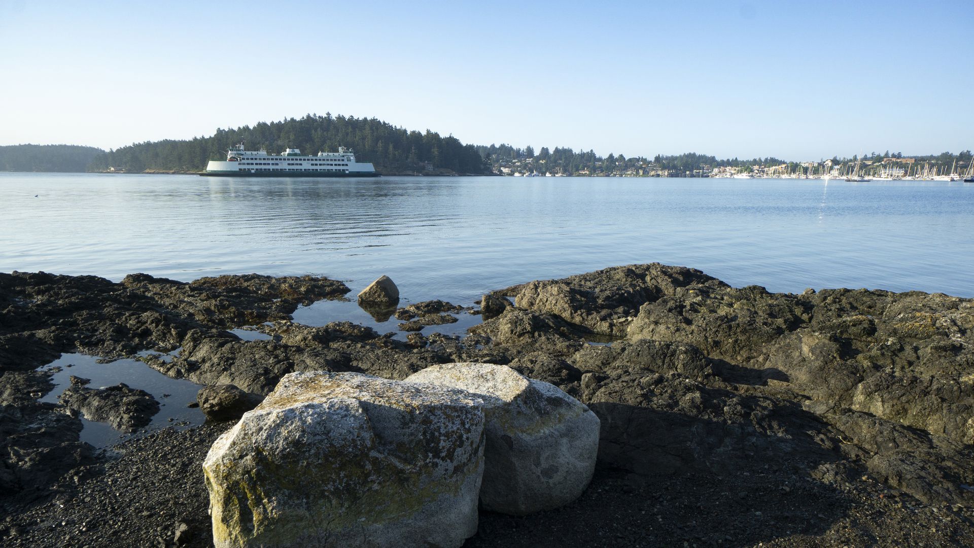 From the shores of a rocky beach, a ferry can be seen sailing into a little port. 