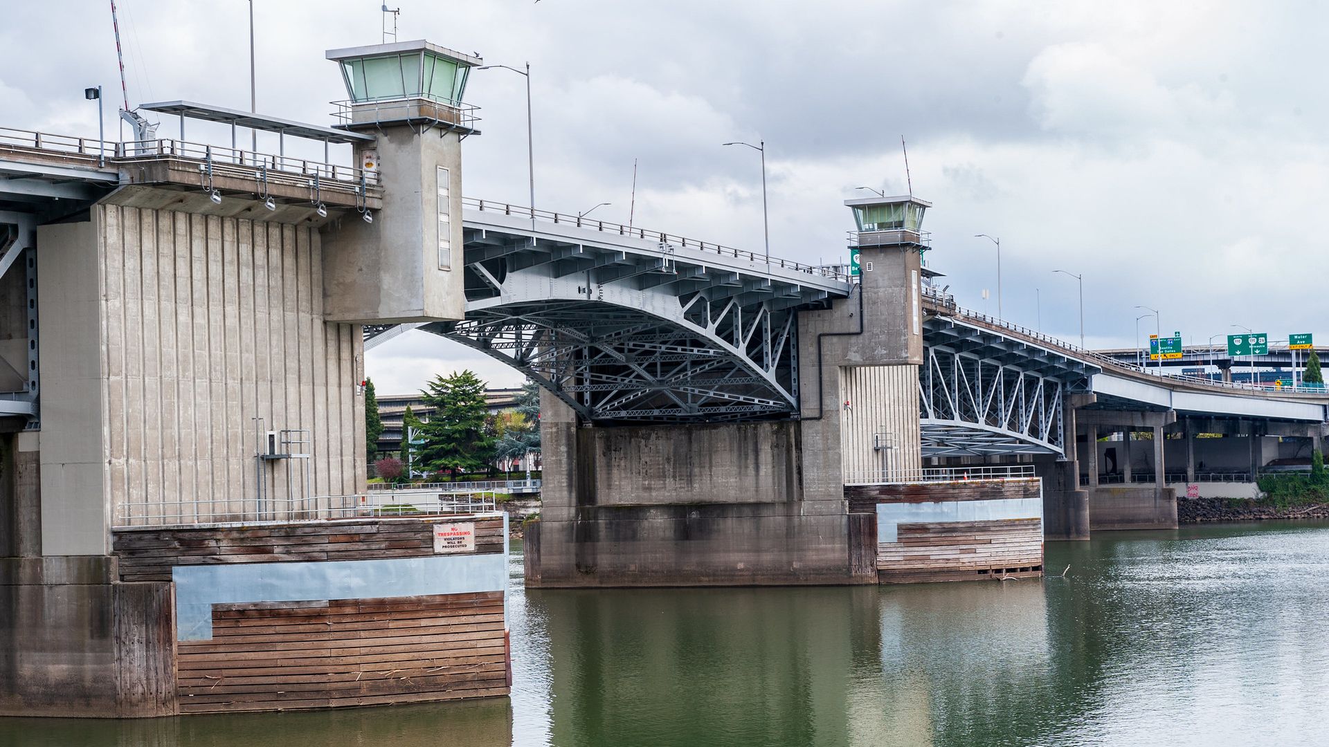 Gray steel and concrete bridge with control towers on each side spanning over calm water under a cloudy sky, with green trees and road signs in the background.