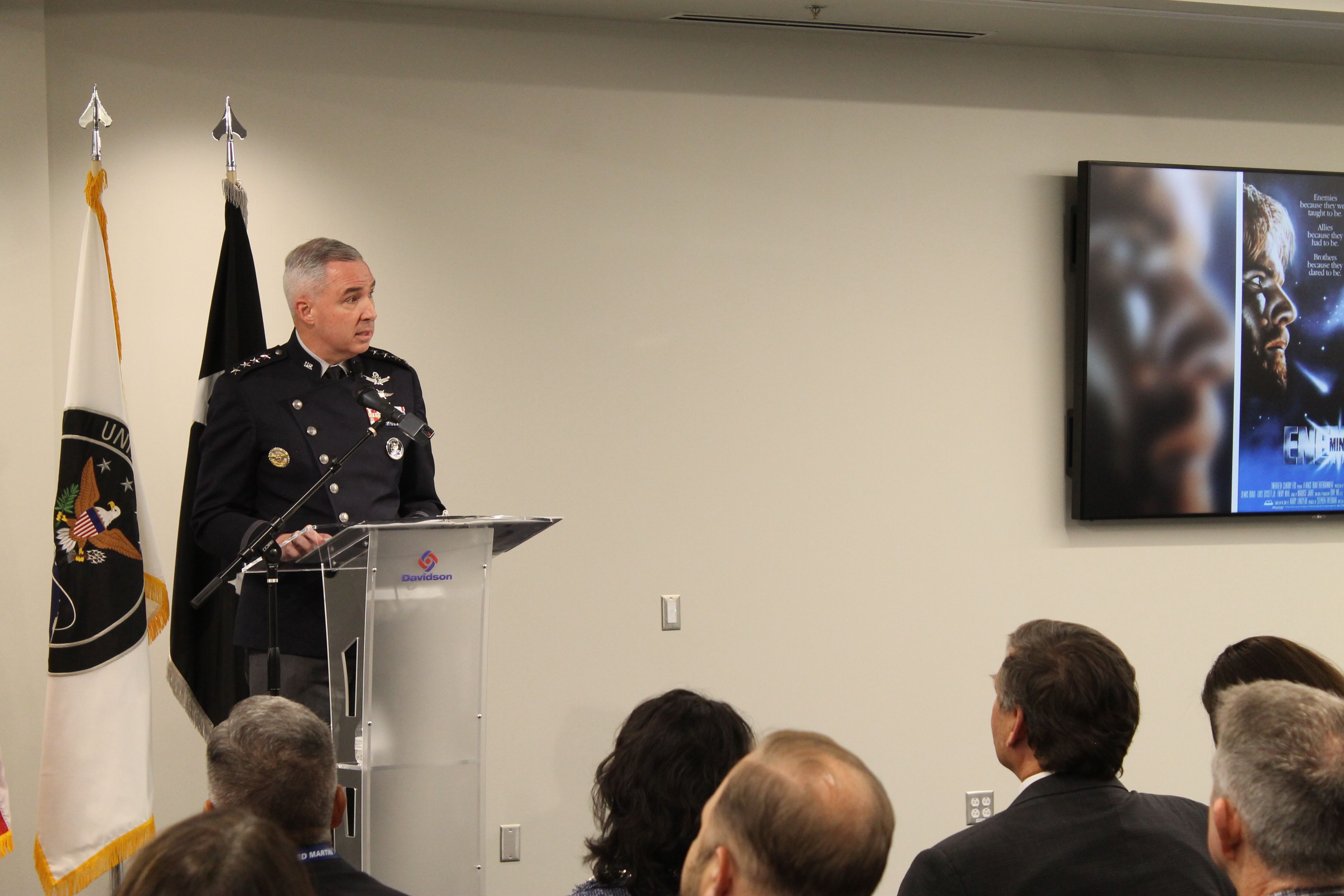 Uniformed military officer speaks at a clear podium in a conference room, flanked by two flags. An audience sits listening as a large screen on the right shows a poster.