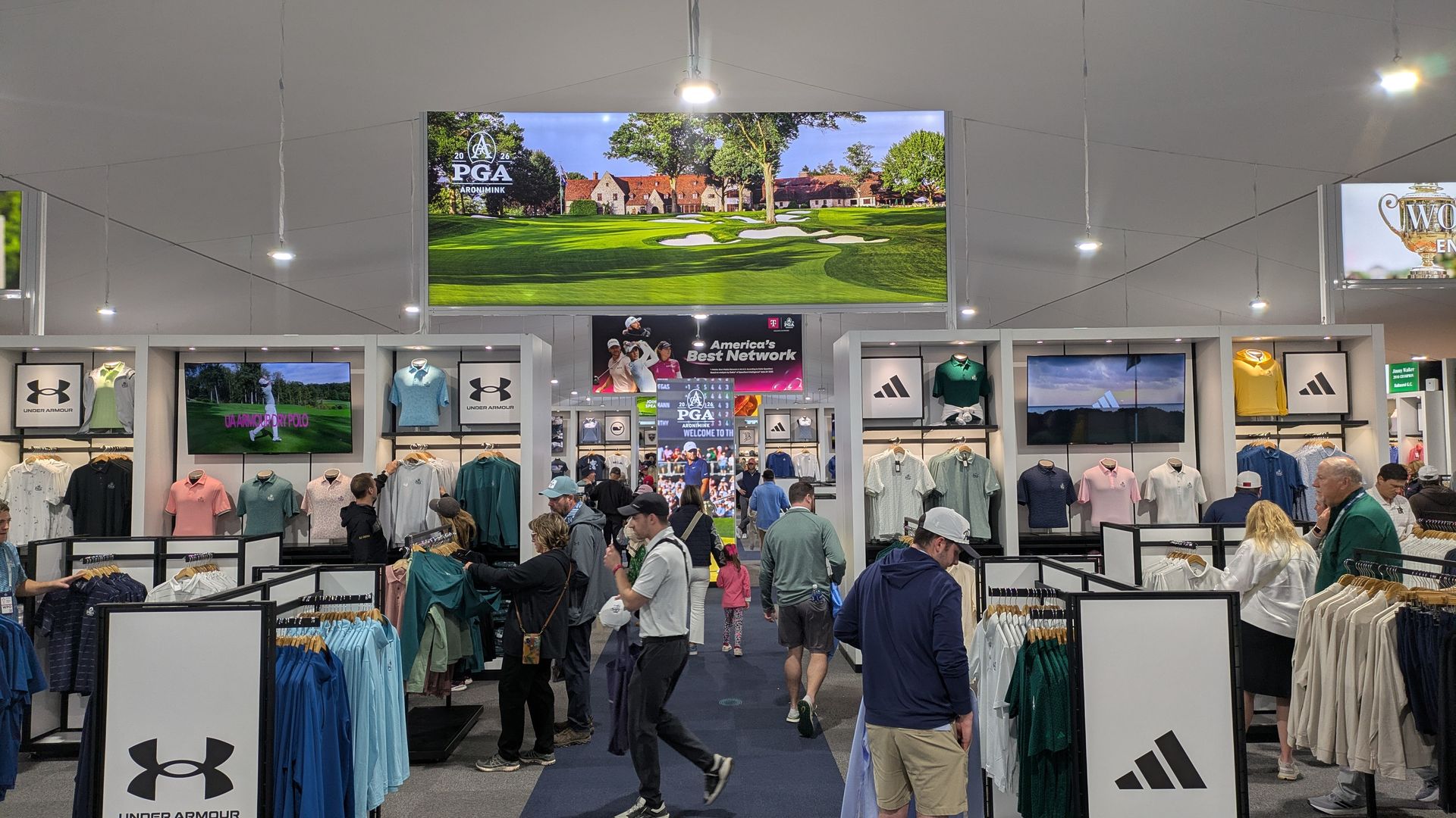 Indoor trade show with Under Armour booths; shoppers browse polo shirts and jackets as a large PGA golf course image dominates a central screen above the displays.