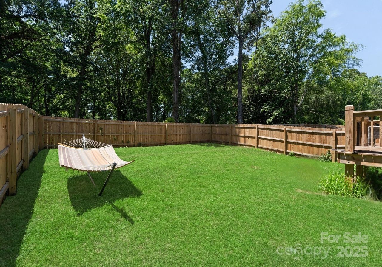 Green grassy backyard enclosed by a wooden fence with tall trees in the background and a beige striped hammock on a metal stand casting a shadow on the lawn.