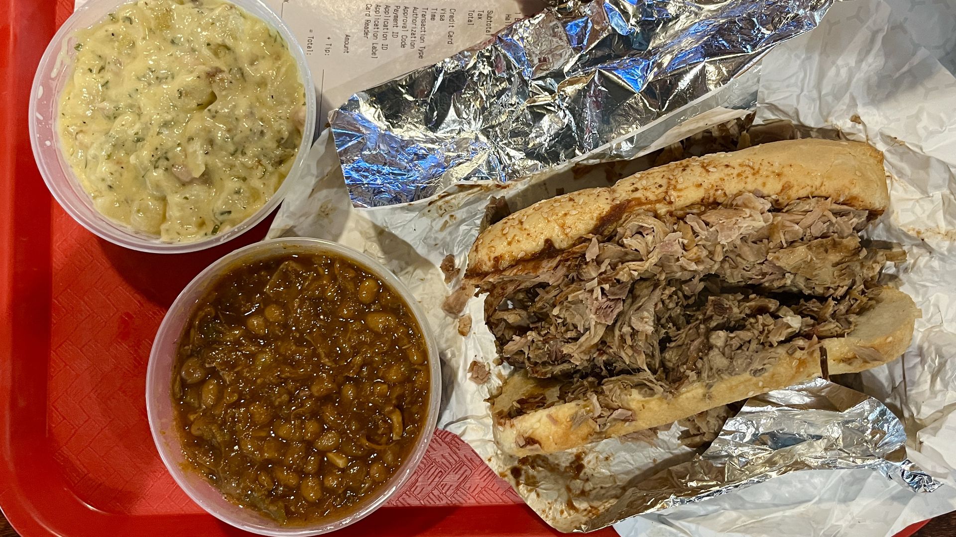 Pulled pork sandwich on sesame bun with two sides: creamy potato salad and baked beans, all placed on a red tray with aluminum and white wrapping and a receipt visible.