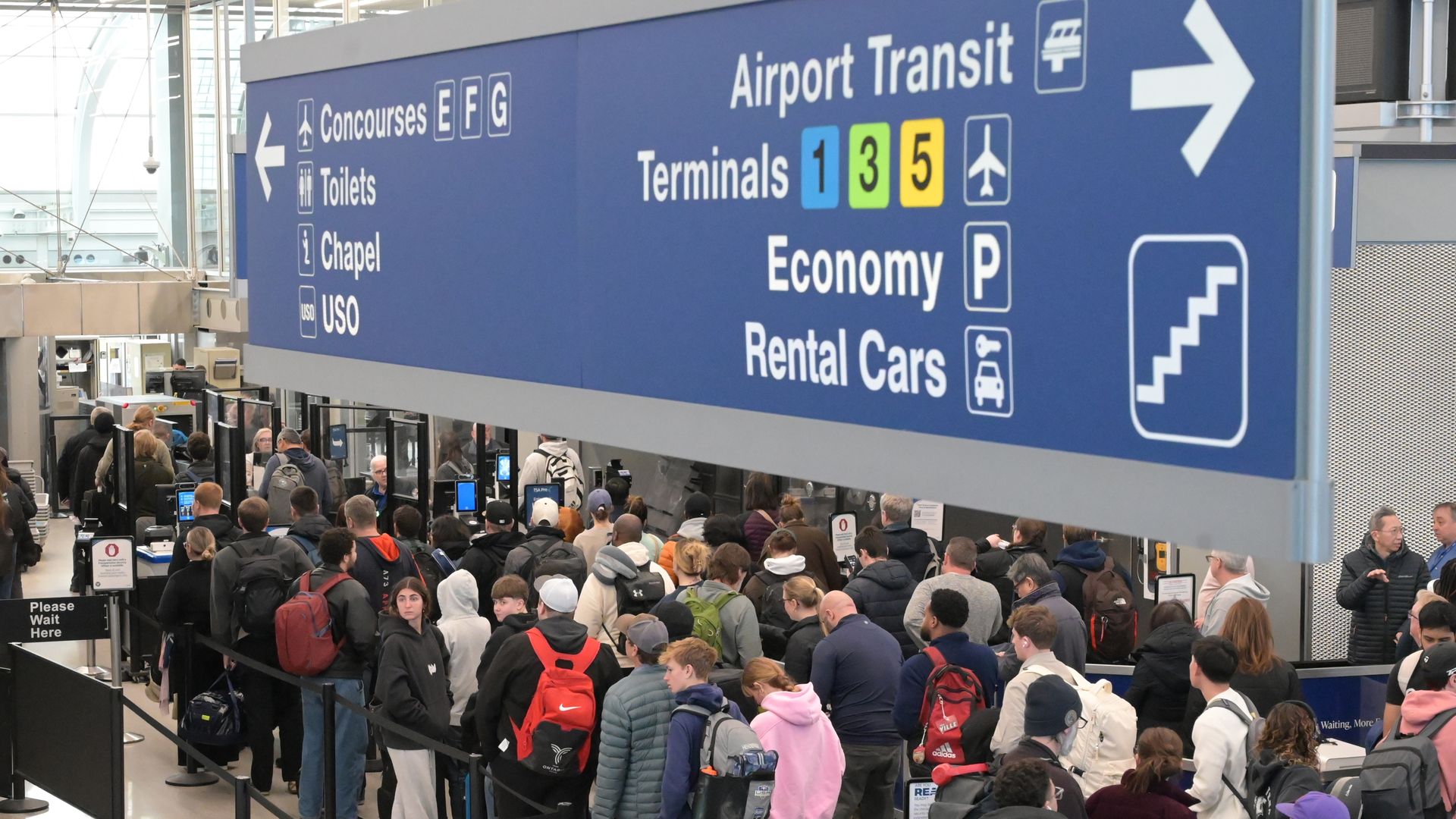 Crowded airport terminal with a long queue of travelers beneath a large blue overhead sign reading "Airport Transit", "Terminals 1 3 5", and "Economy Rental Cars"; many carry backpacks and luggage.