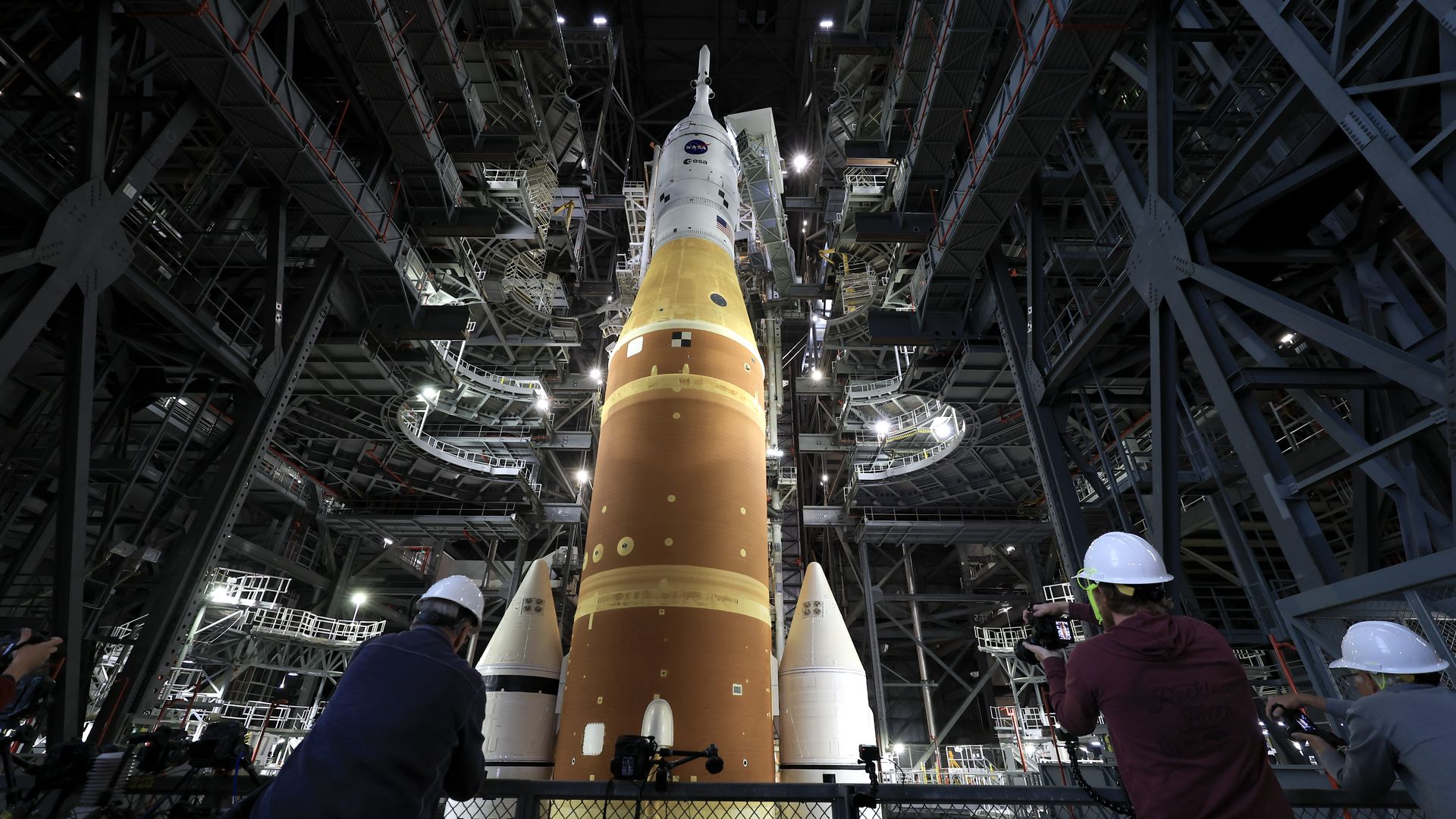 Large NASA rocket inside a massive metal launch structure, with three people wearing white hard hats photographing it from behind. The rocket's lower part is orange and upper part white.