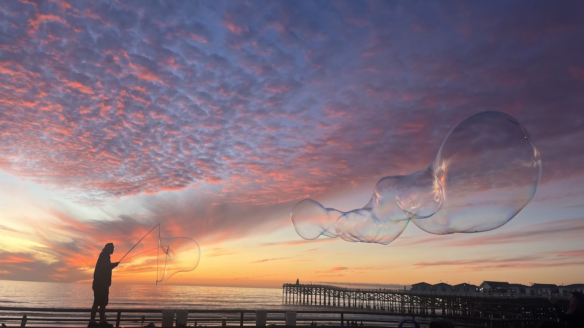 A man makes large bubbles in front of a pink and orange sunset at Pacific Beach
