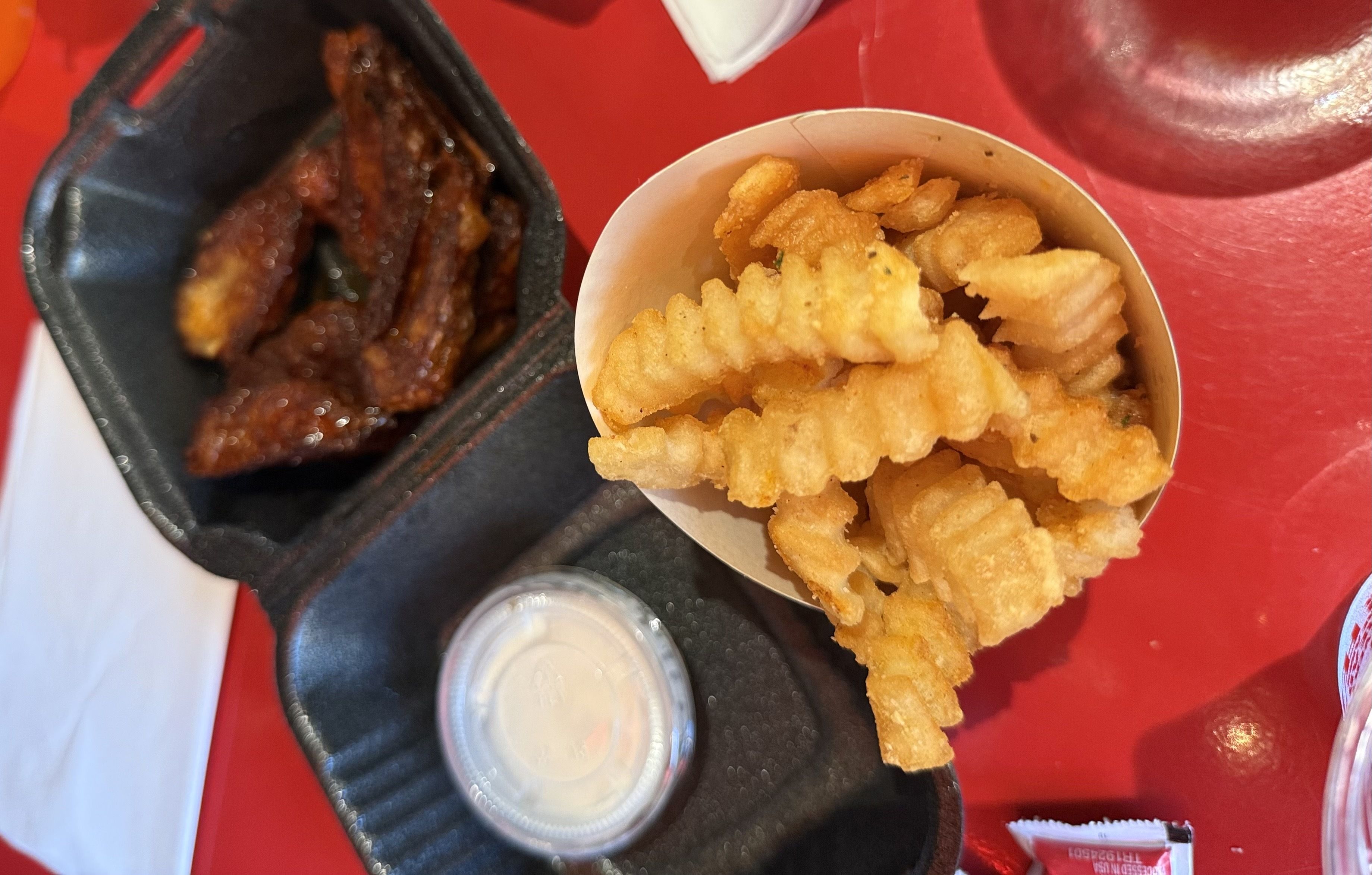 Pile of golden, crinkle-cut french fries in a white paper cup on a red table, with a dark tray of glazed ribs beside them, and a small cup and napkin nearby.
