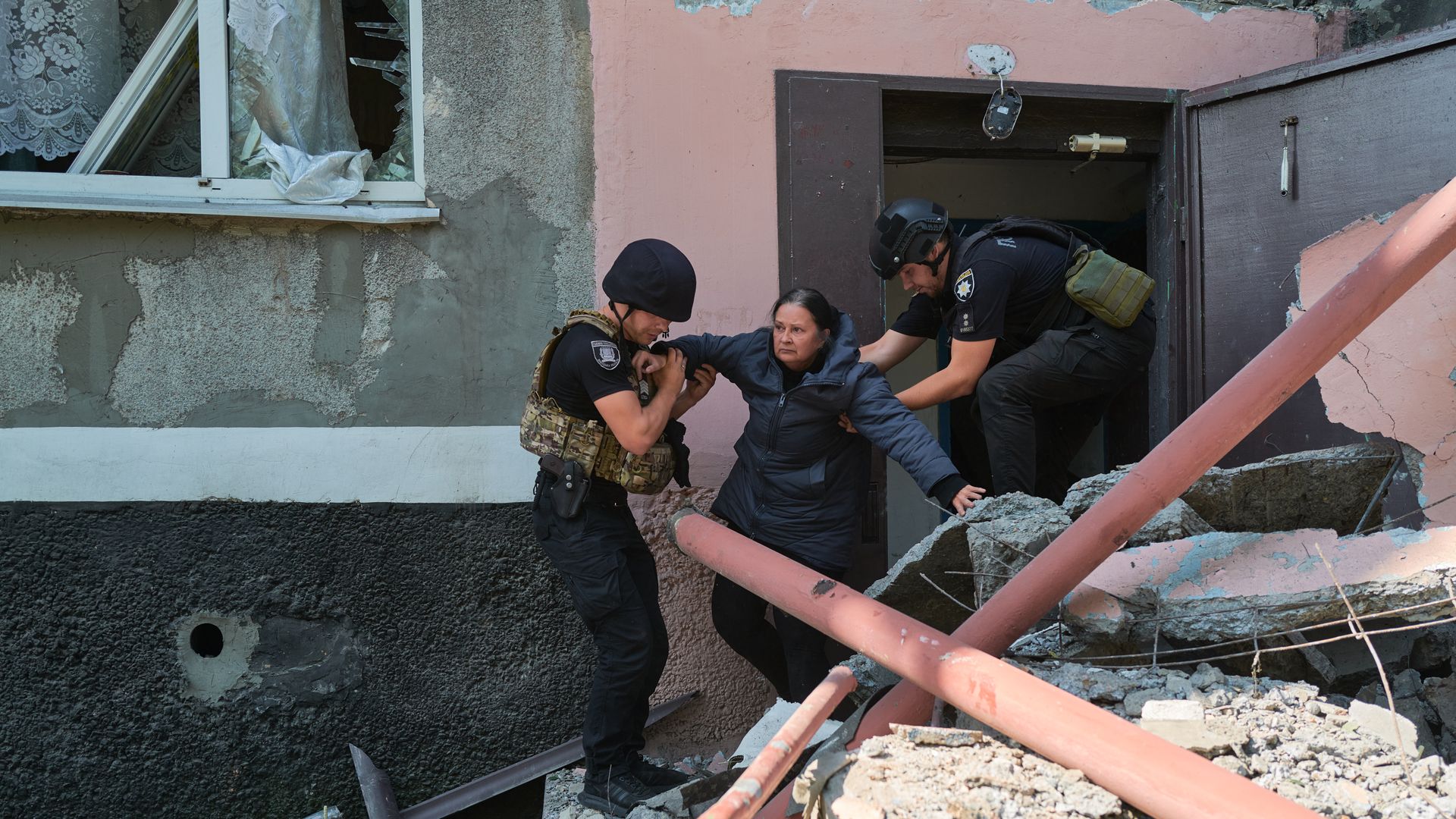 Two armed police officers help a woman in a blue jacket out of a damaged building with broken window and rubble around, indicating a disaster or conflict zone.